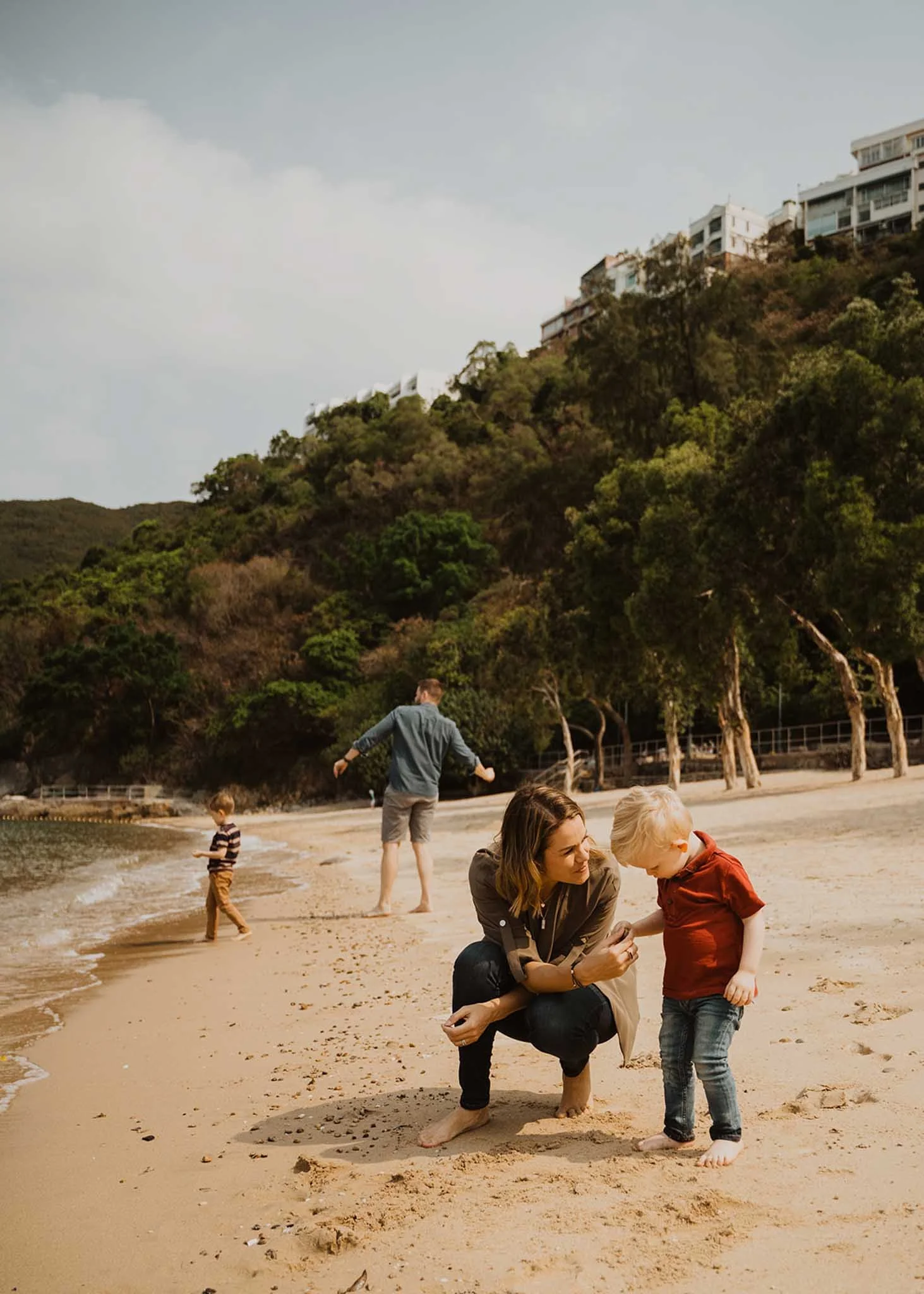 A woman helping a young boy on a sandy beach, with other children playing near the water, and a hillside with houses in the background.