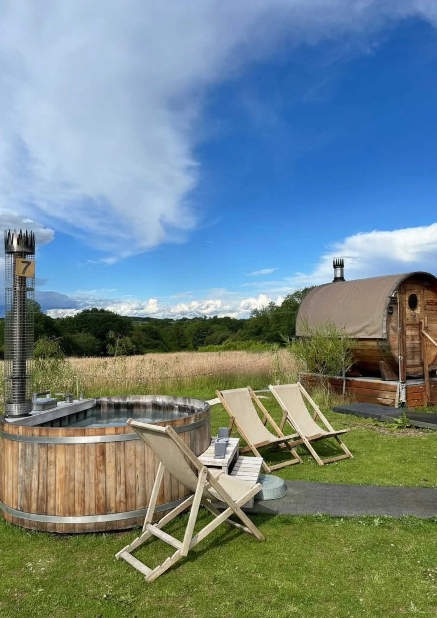 Outdoor scene with a hot tub, wooden chairs, a wooden shed, and a field of grasses under a partly cloudy sky.