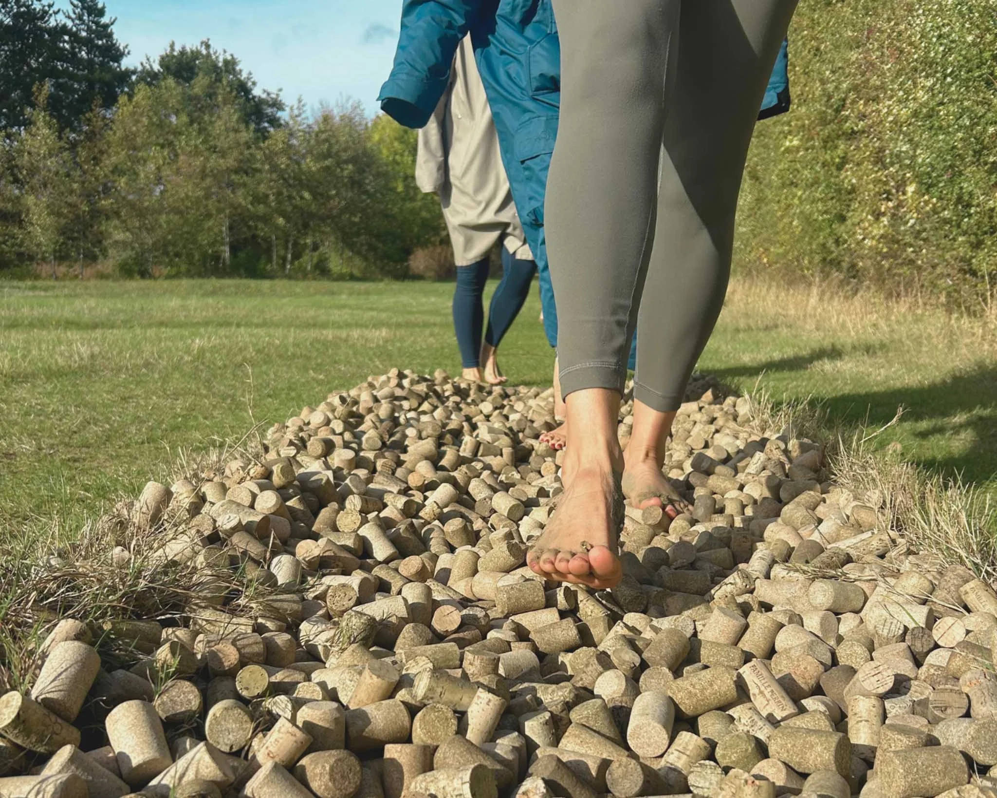 People walking barefoot on a pile of small rocks outdoors with trees in the background.