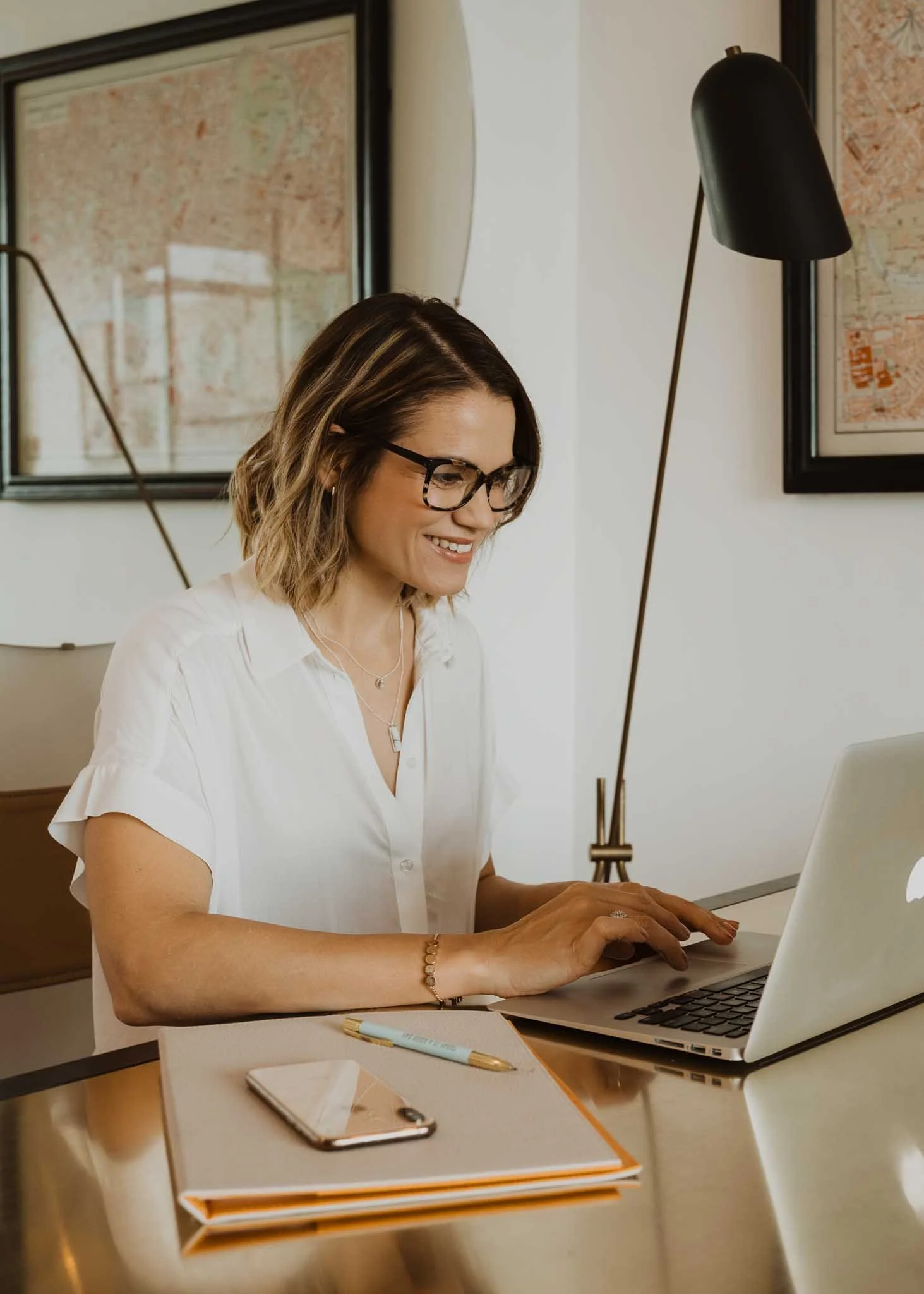 A woman with glasses sitting at a desk working on a laptop, smiling. The desk has a notebook, a tablet, a pen, and a smartphone. The background has framed maps on the wall, and there is a black floor lamp with a gold stand.