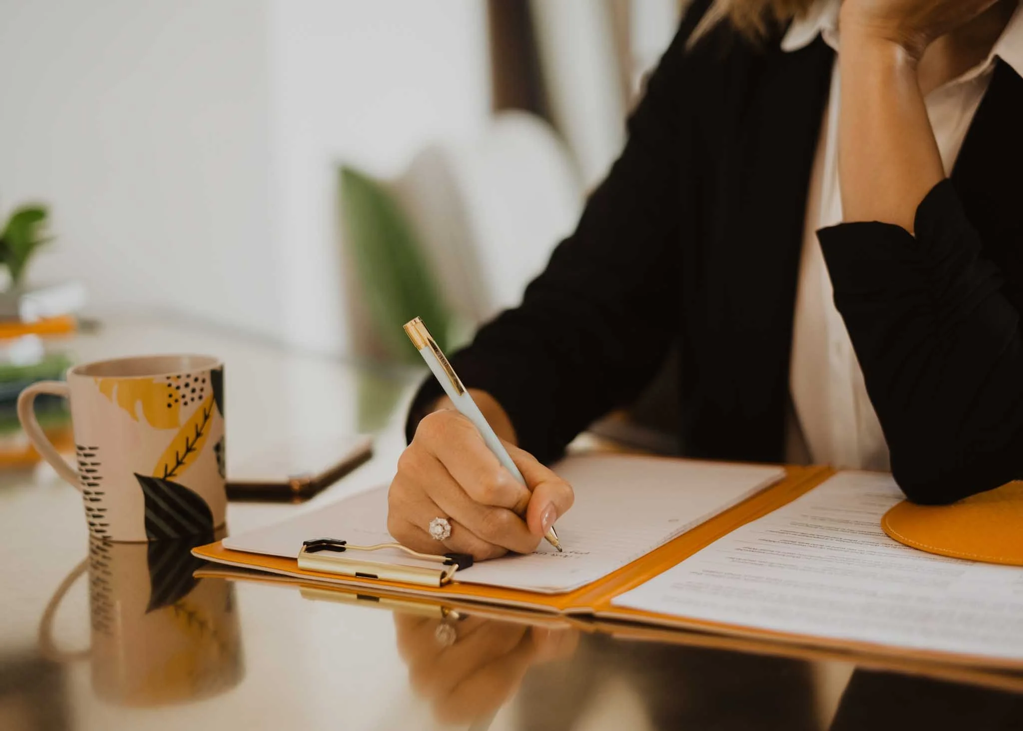 A person sitting at a desk, writing in a notebook with a pen. There is a mug with black and yellow patterns, some papers, and a small plant in the background.
