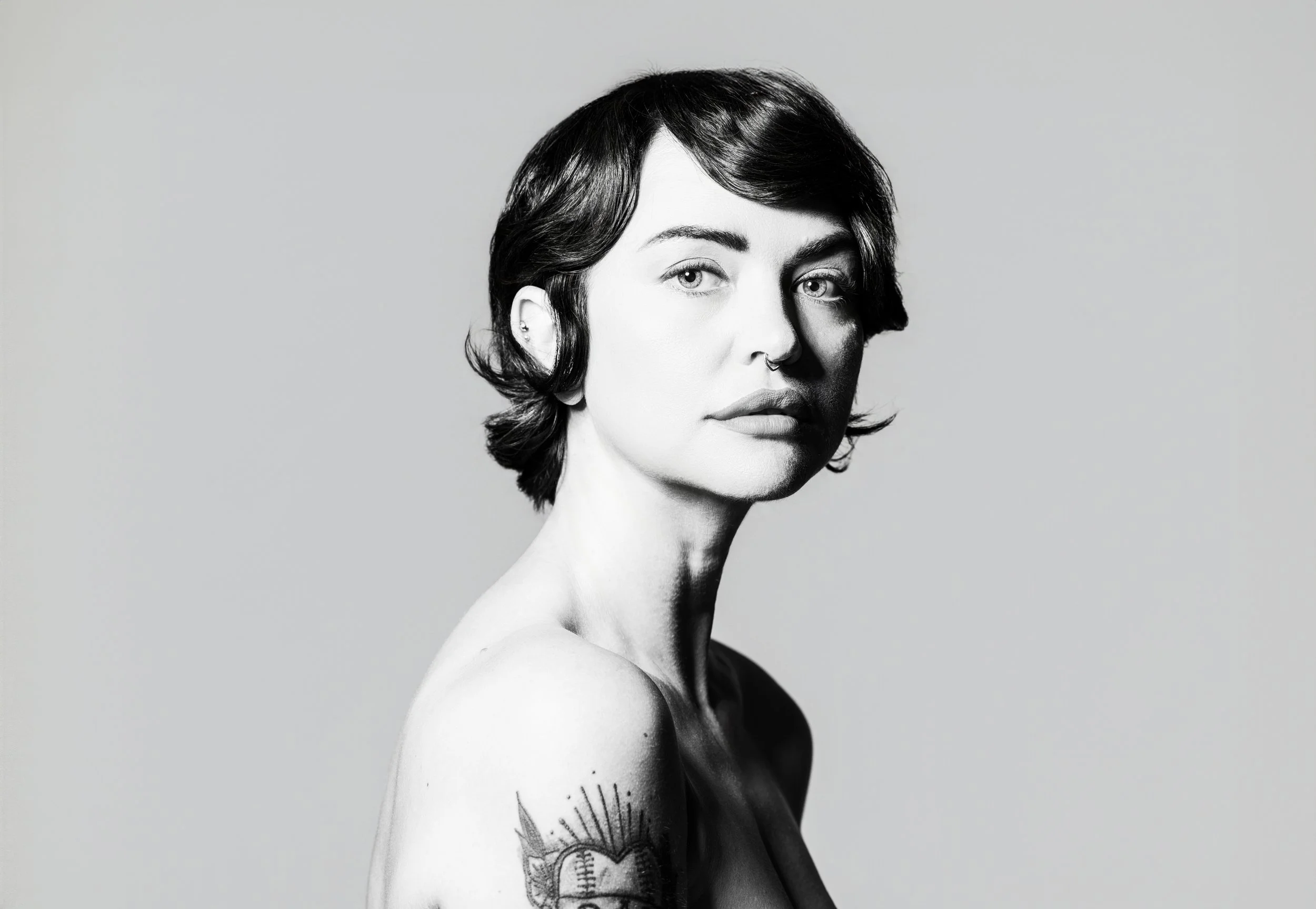 Black-and-white portrait of a woman with short dark hair, tattoos on her left arm, and a septum piercing, looking at the camera against a plain background.
