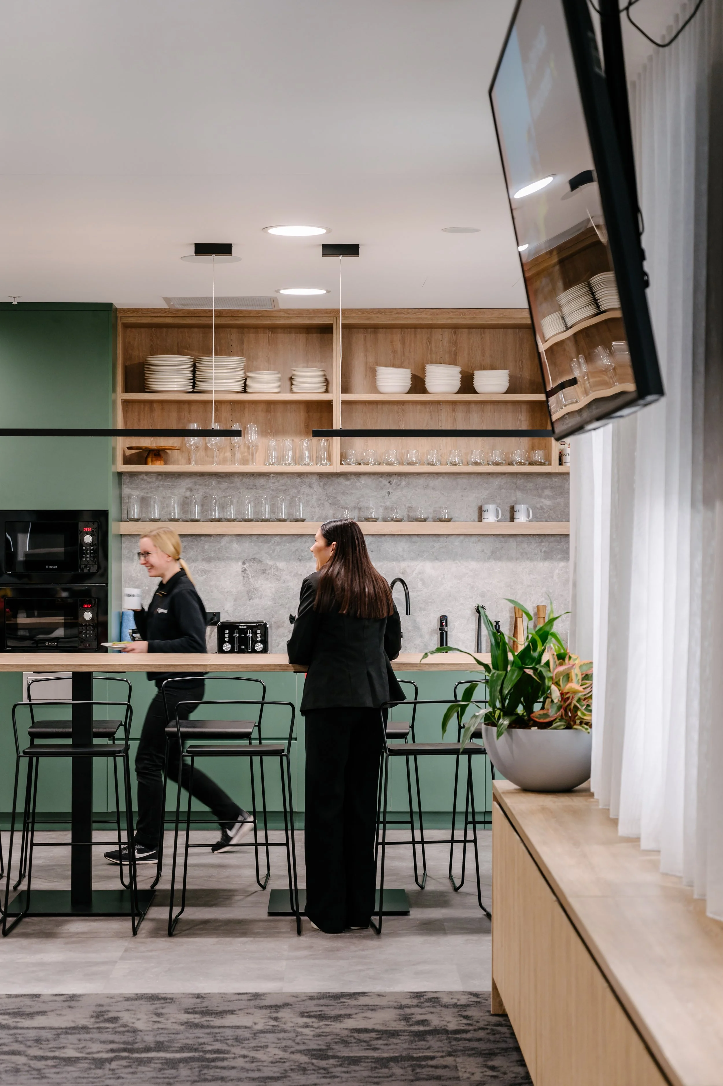 Two women working in a modern kitchen with wooden shelves holding white dishes and glasses, a black countertop, green cabinets, and a large flat-screen TV mounted on the wall.