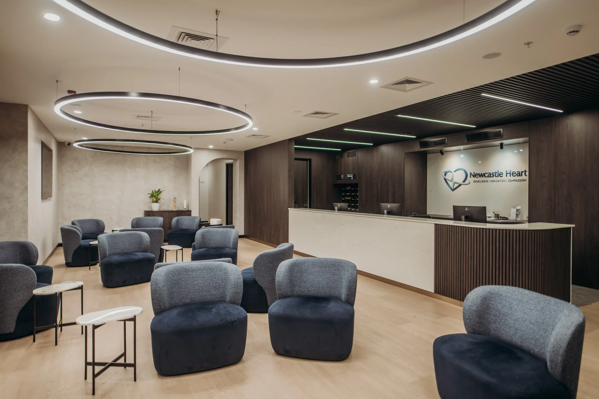Modern waiting area or lounge with multiple gray upholstered chairs arranged in clusters, small side tables, a reception desk with the logo 'Newcastle Heart,' and ceiling lights with circular and linear LED fixtures.