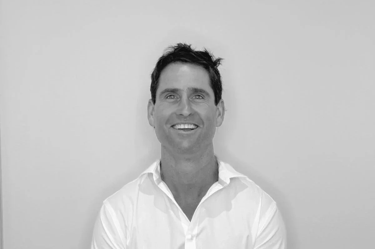 Black and white photo of a smiling man with short dark hair, wearing a white dress shirt, standing against a plain wall.