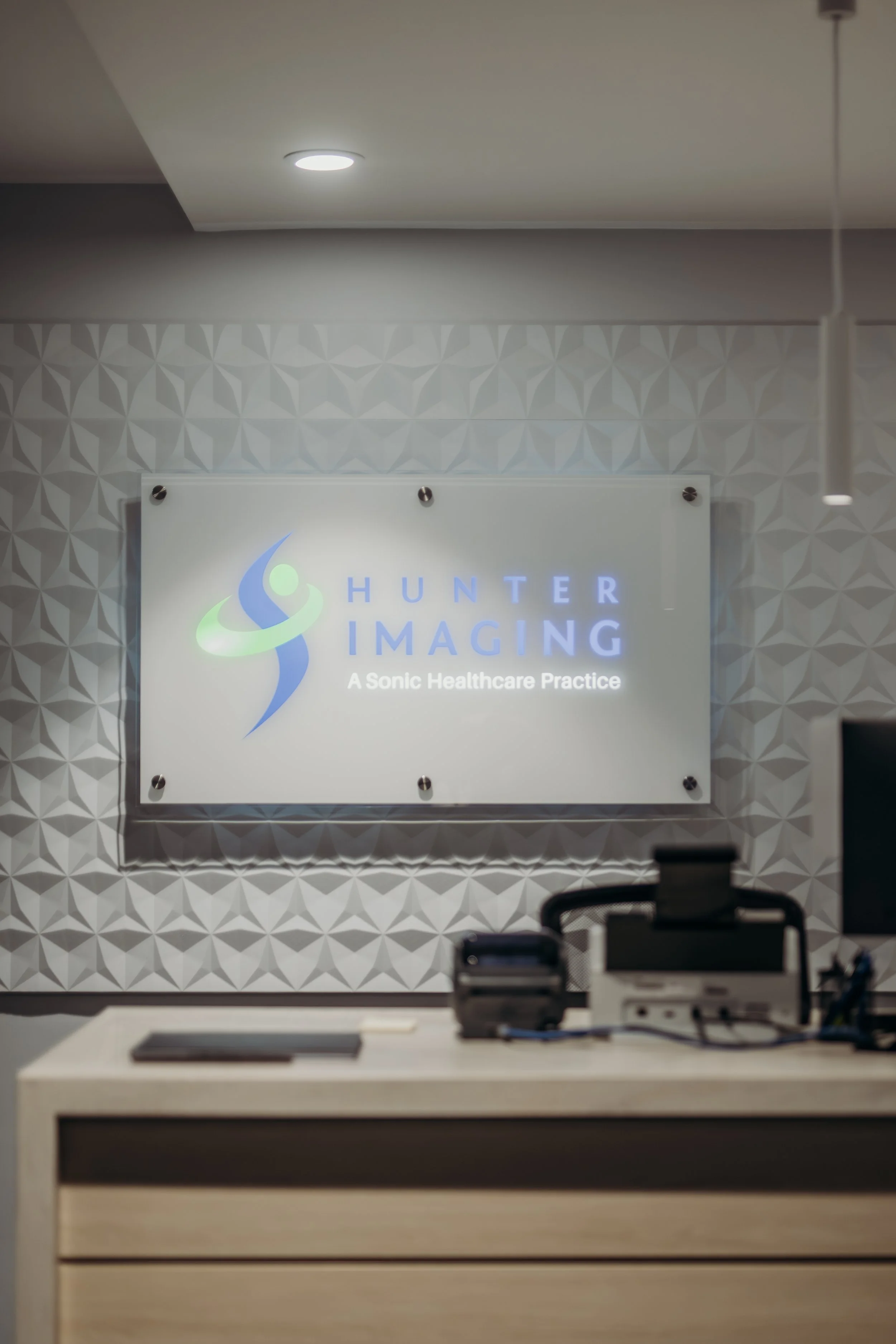 Interior of an office or reception area displaying a sign for Hunter Imaging, a Sonic Healthcare practice, mounted on a wall with a geometric patterned background. Desk or counter with some office equipment in the foreground.