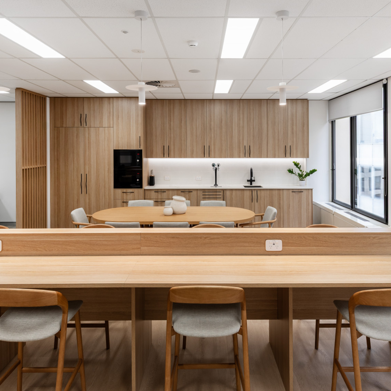 Modern kitchen and dining area with wooden cabinets, round wooden table, and chairs, large window with blinds, and minimal decor.