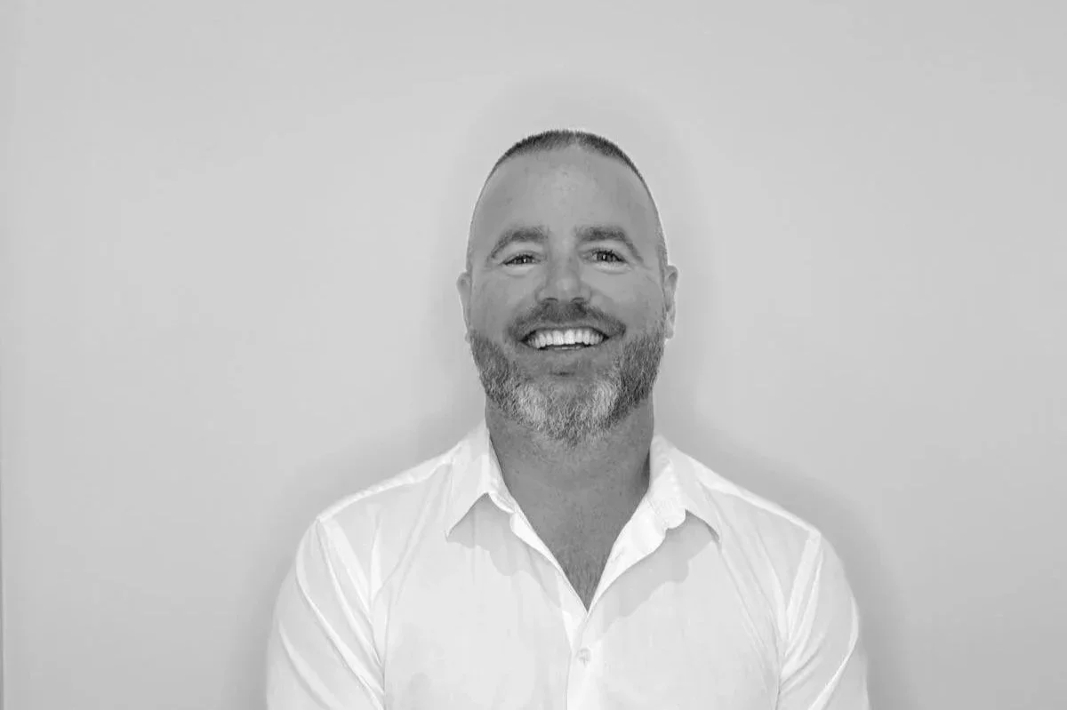 A smiling man with short hair and a beard, wearing a white shirt, standing against a plain wall.