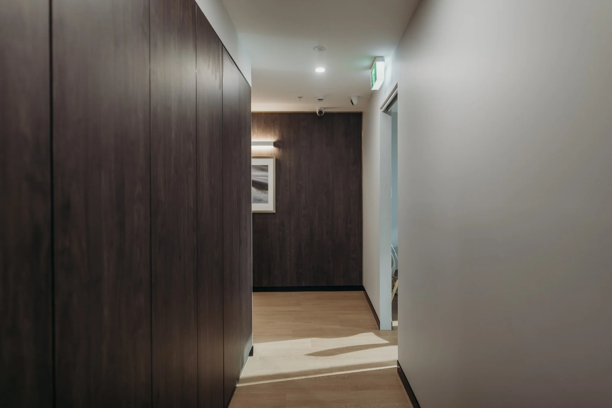 A hallway in a modern building with a dark wooden wall on the left, light-colored walls on the right, and a picture frame at the end of the corridor. There is a ceiling light and an illuminated exit sign above the doorway on the right.