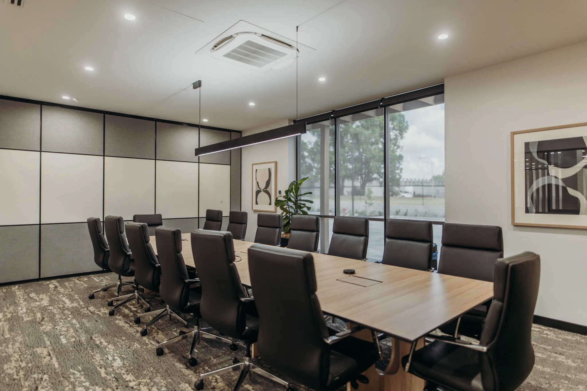 A conference room with a long wooden table surrounded by black leather office chairs. Large windows with blinds allow natural light, and there are framed abstract artworks on the wall, with a view of trees and a parking lot outside.