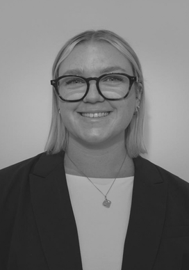 A woman smiling, wearing glasses, a blazer, and a necklace, in black and white portrait.