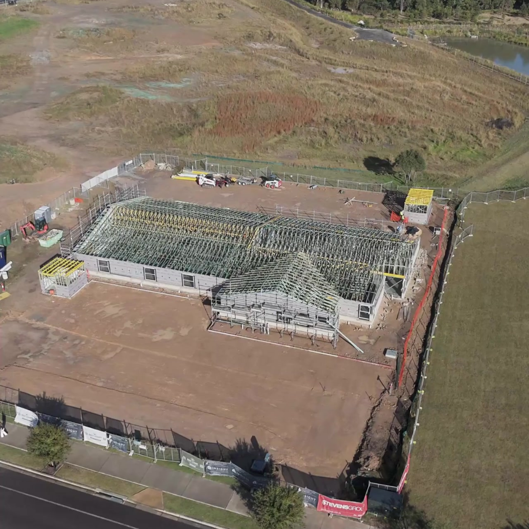 A house under construction with a partially completed roof framing in a fenced area, surrounded by open land and a road in the foreground.