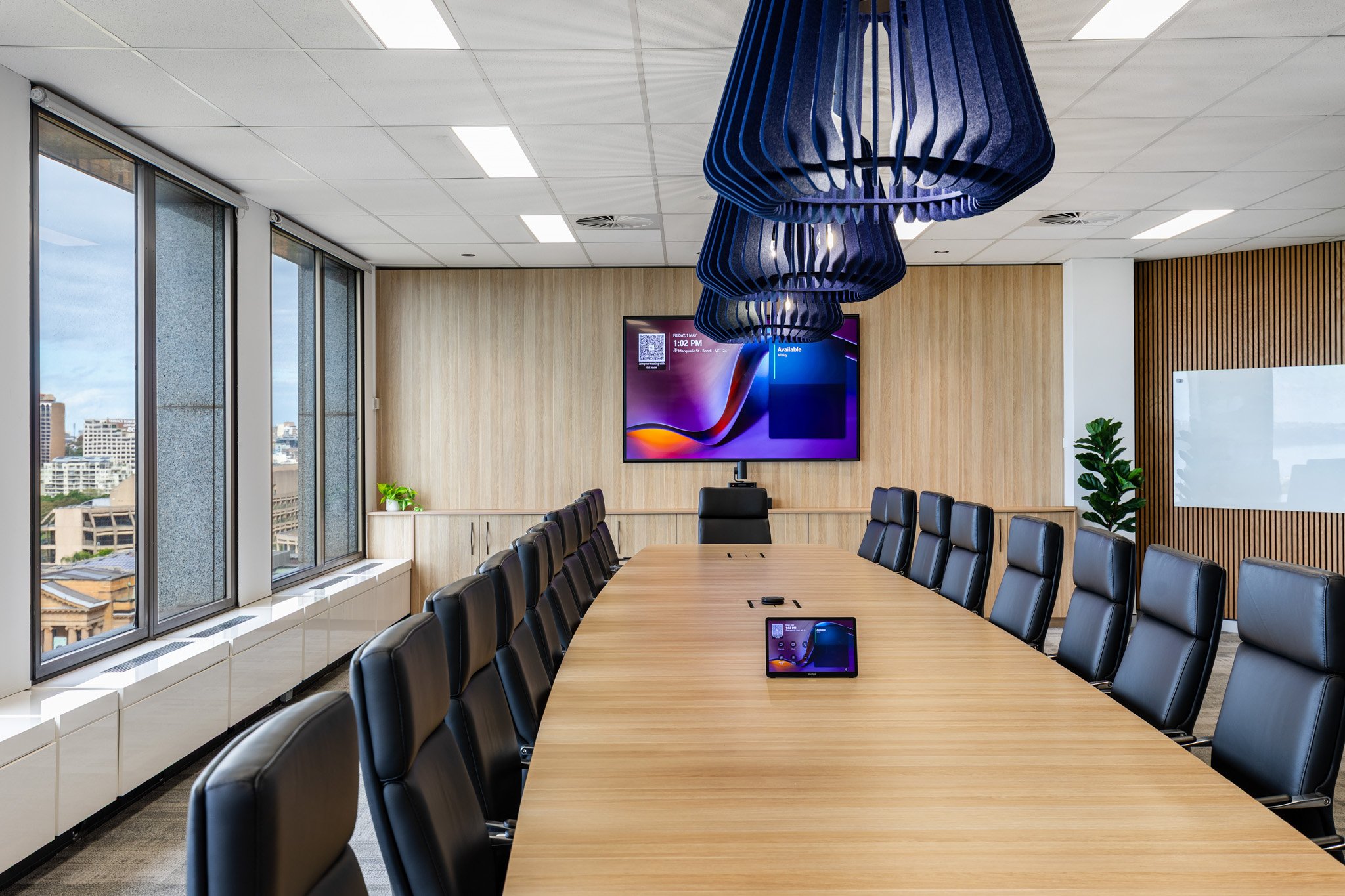 A modern conference room with a long wooden table, black leather chairs, large windows with city views, a digital display on the wall, and decorative blue light fixtures hanging from the ceiling.