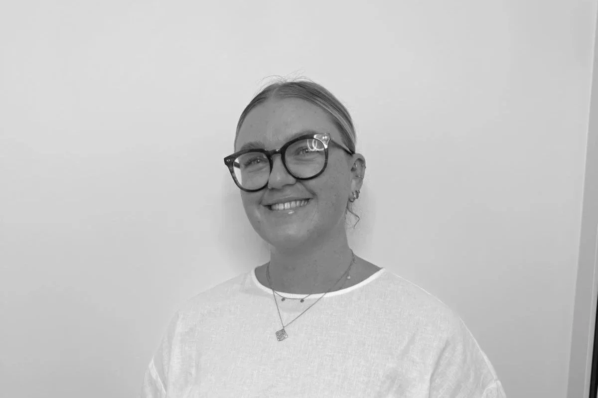 Black and white photo of a woman with glasses and earrings, smiling, wearing a white shirt and layered necklaces, standing against a plain wall.