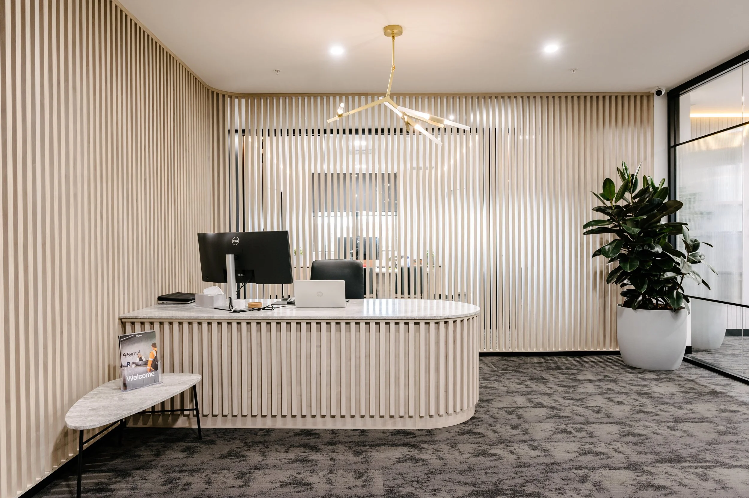 Modern office reception area with a curved wooden slat divider behind a desk, a computer, and a potted plant on the right. A chandelier hangs from the ceiling.