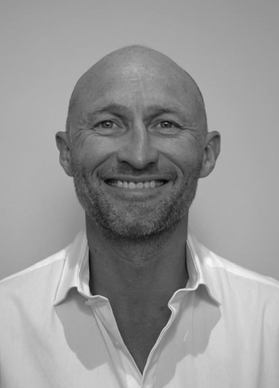 Black and white photo of a smiling bald man with a beard, wearing a white collared shirt.