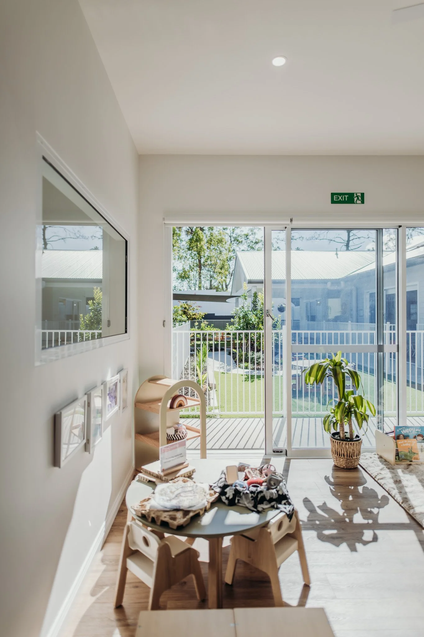 A bright nursery room with children’s furniture, plants, and toys. Large glass doors lead to an outdoor balcony with a white railing, giving a view of neighboring houses and trees.