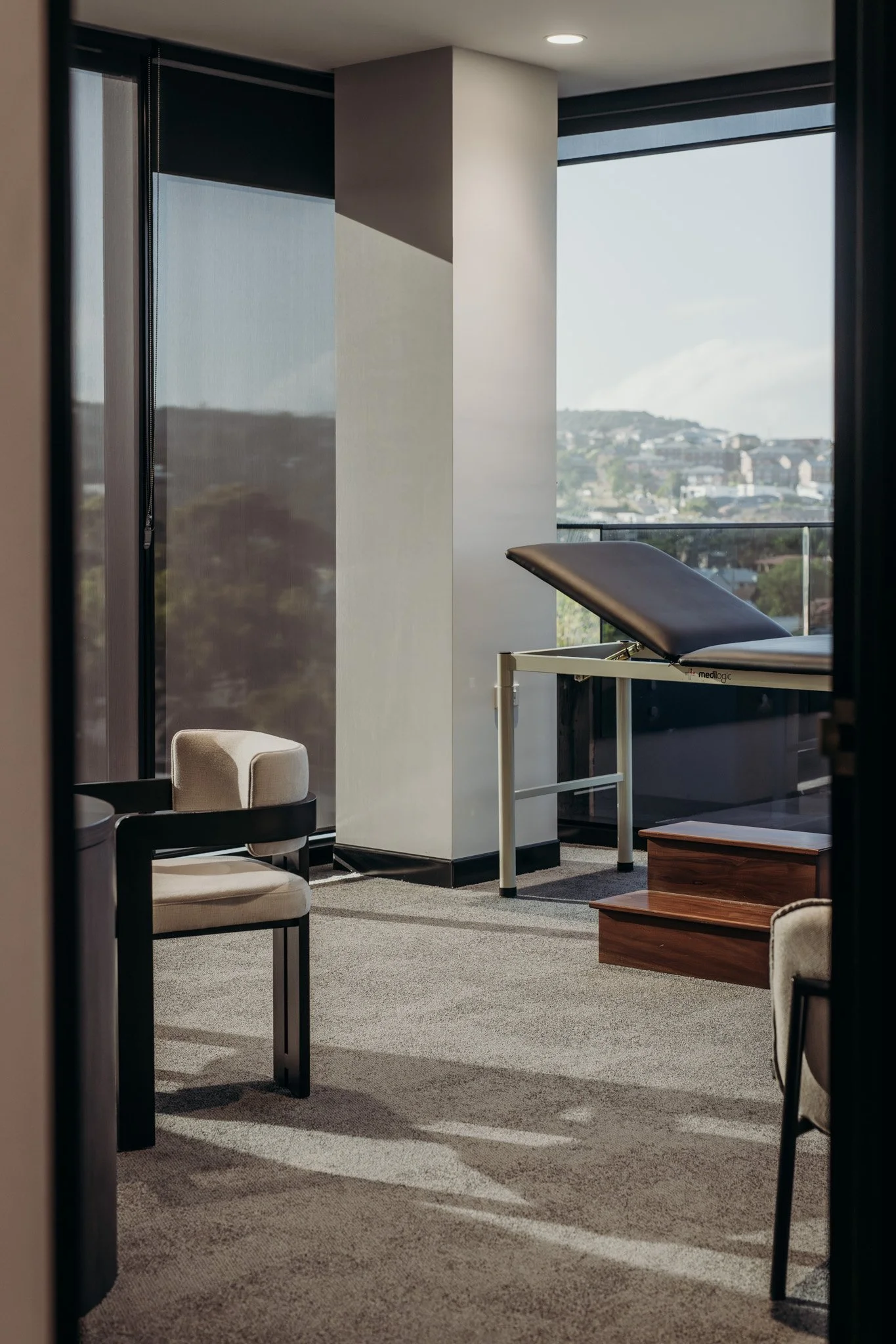 Interior of a medical or massage room with a treatment table, chairs, and large windows showing a city view.