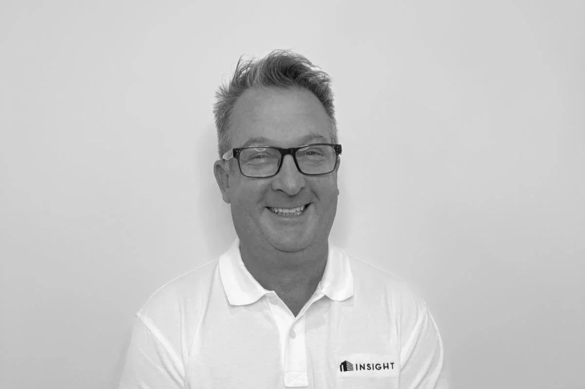 Black and white photo of a smiling man with glasses, wearing a white polo shirt with a logo on the left side that says 'Insight', standing against a plain wall.