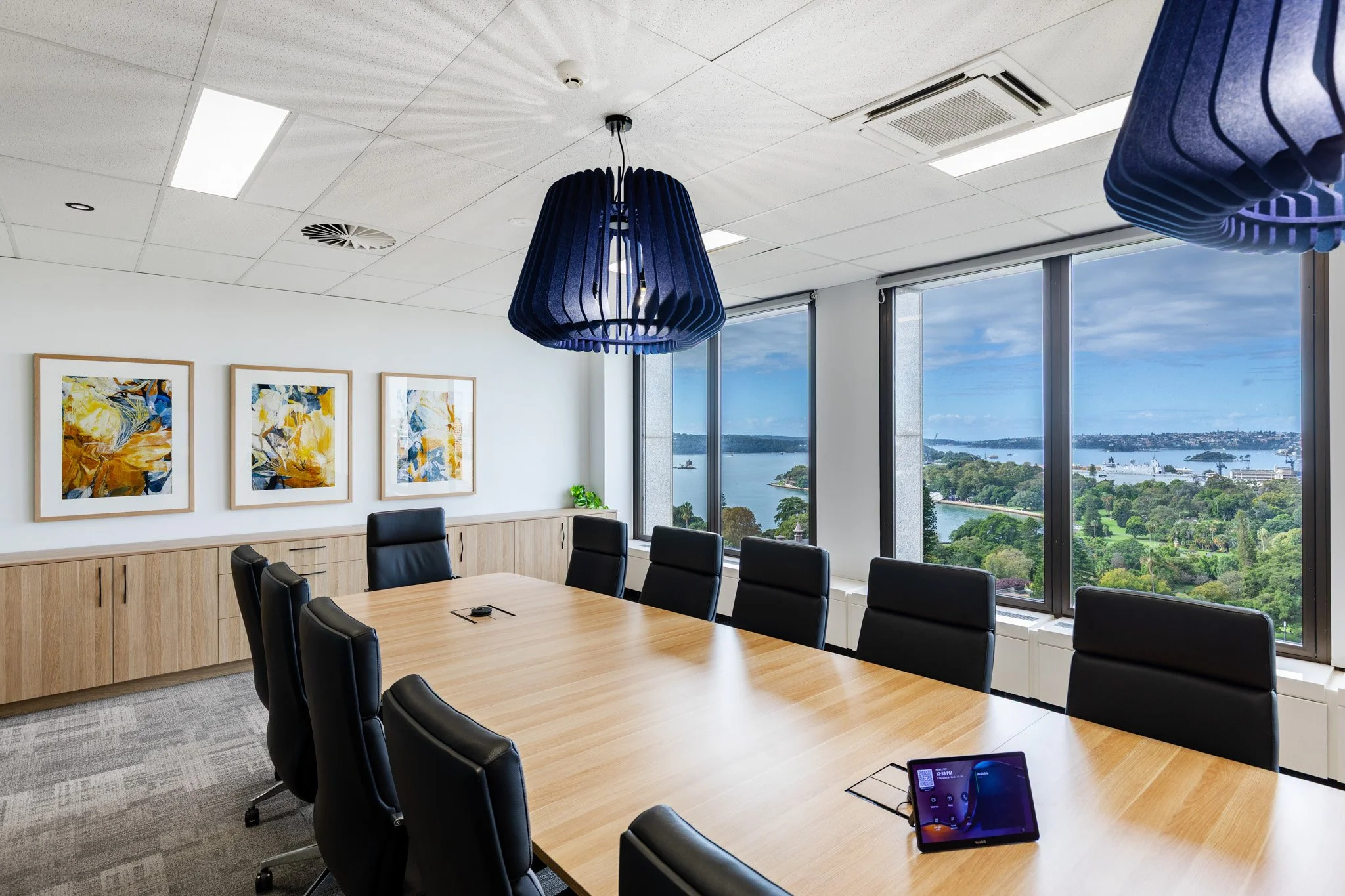 An conference room with a large wooden table, black chairs, and large windows showing a river and trees outside. The room has wall art, ceiling lights, and a hanging blue pendant lamp.