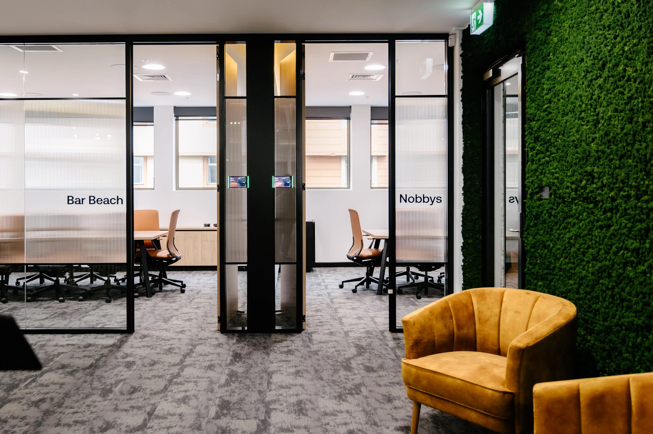 Modern office space with glass partitions, orange chairs, a green moss wall, and a yellow velvet armchair.