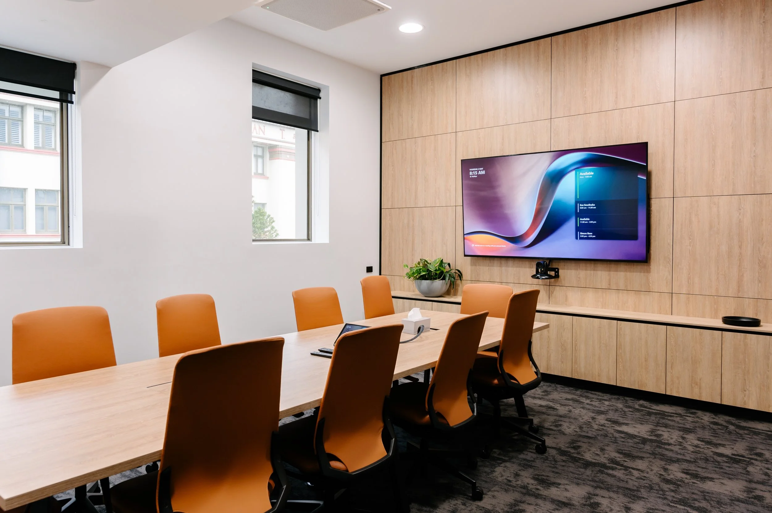 A conference room with a long wooden table surrounded by orange chairs, a large flat-screen TV on a wall with wood paneling, a potted plant, windows with black blinds, and office supplies on the table.