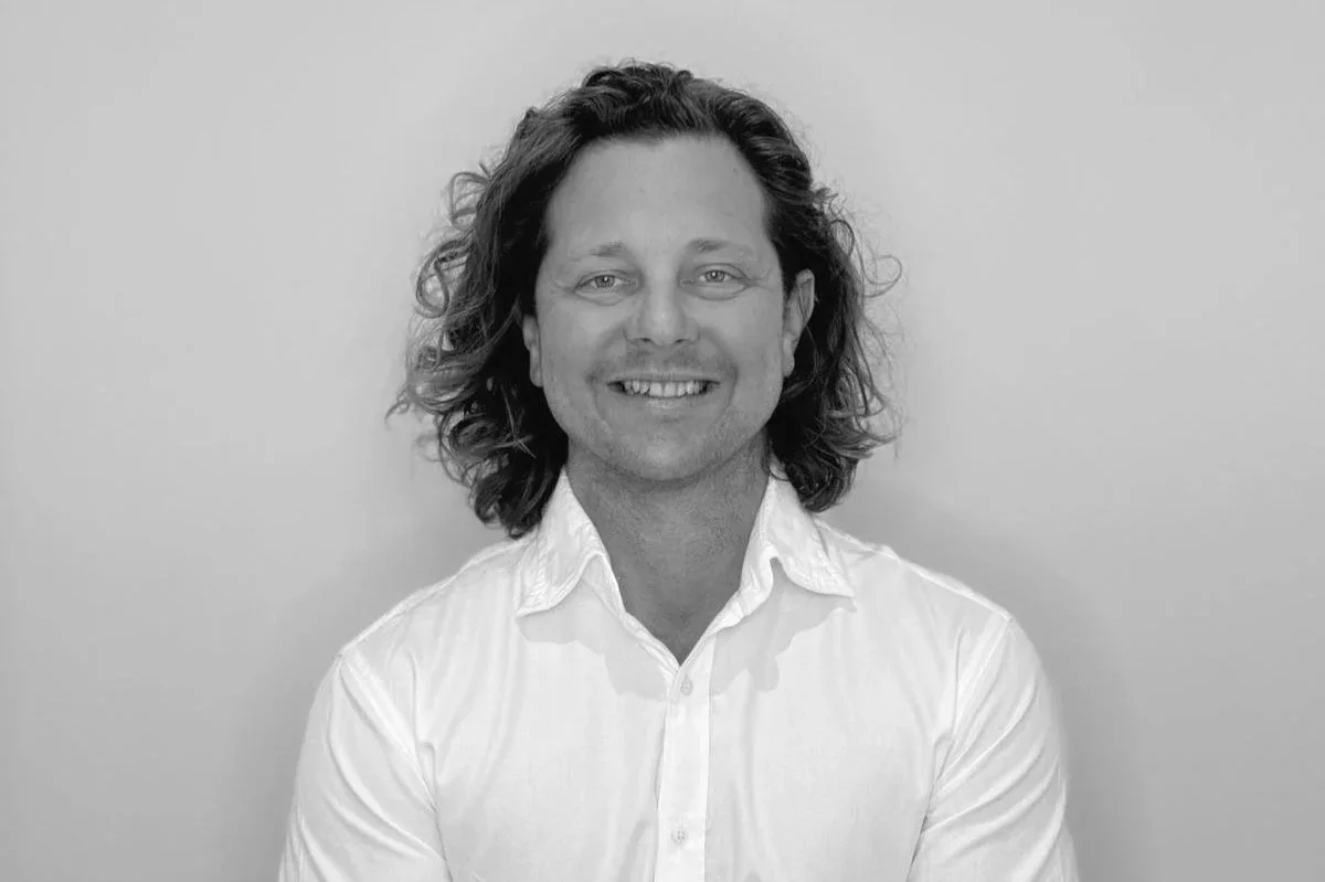 Black and white portrait of a smiling man with long, wavy hair wearing a white button-up shirt against a plain background.