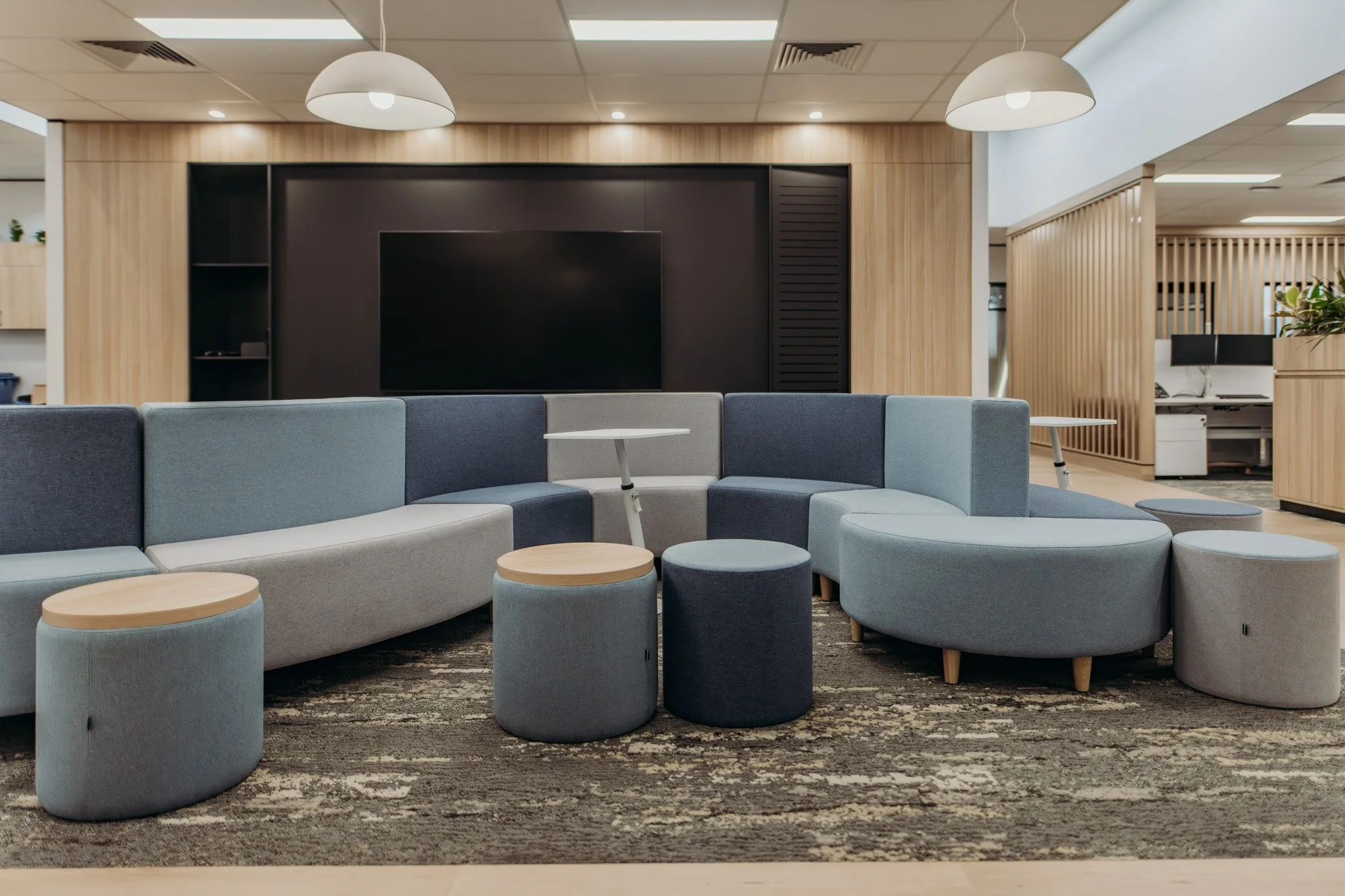 Modern office lounge area with a cozy, curved sectional sofa in shades of blue and gray, surrounded by small round ottomans, with a large flat-screen TV on a wooden accent wall and contemporary pendant lighting.