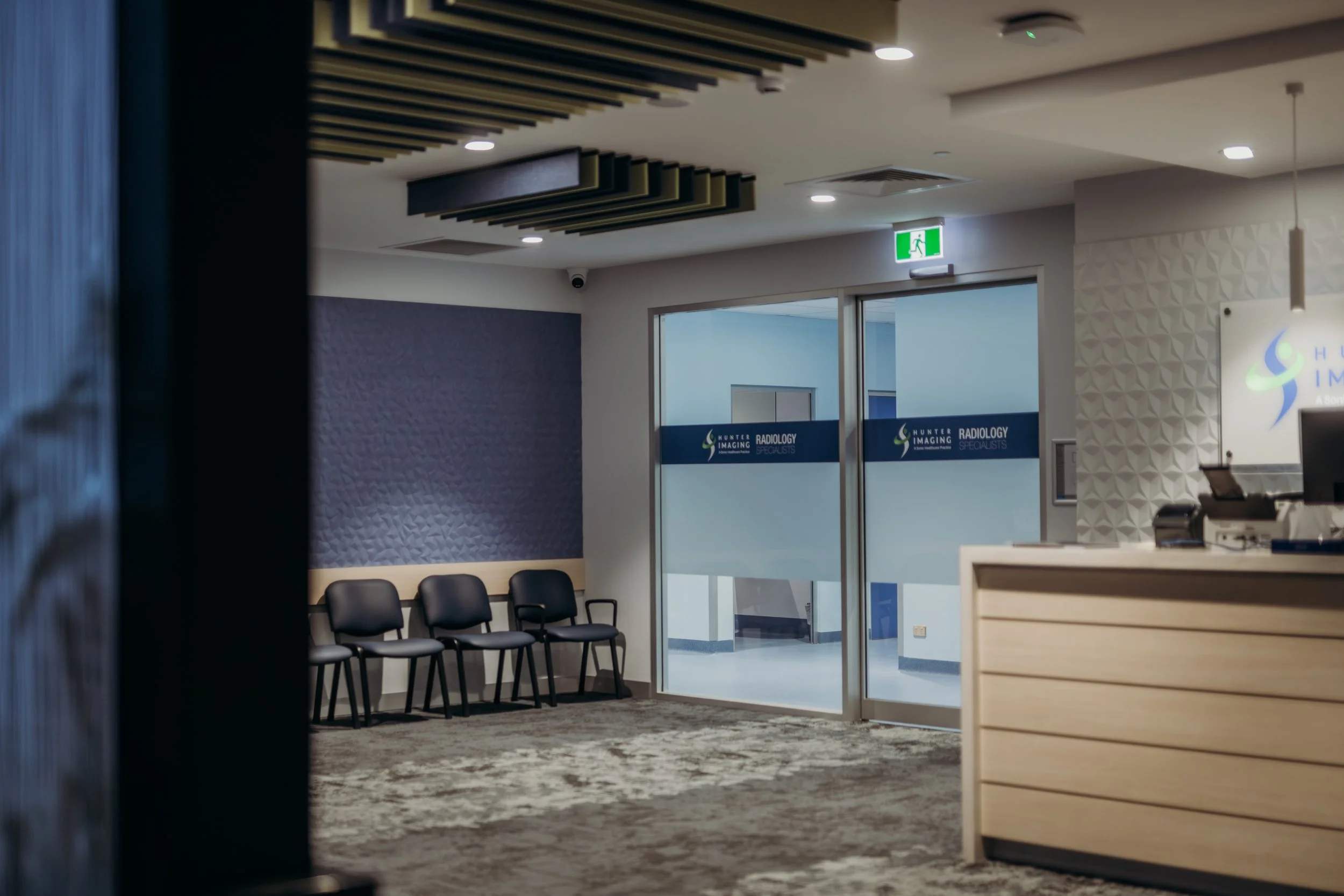 Interior view of a medical office or clinic waiting area with black chairs, a glass door with hospital signage, and a reception desk. Exit sign is visible above the door.