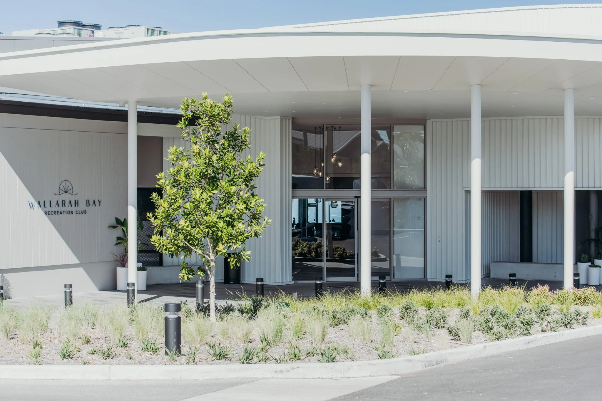 Exterior view of the Wallarah Bay Recreation Club, a modern building with white paneling, large glass windows, and a flat overhang supported by white columns. There are trees and landscaping in the foreground, with a parking lot curb and lighting posts along the street.