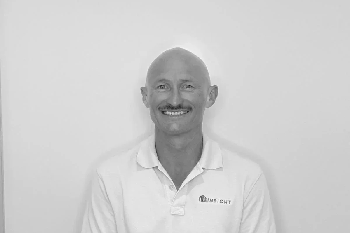 A smiling man with a shaved head and mustache, wearing a white polo shirt with a logo that says 'INSIGHT', standing against a plain wall.