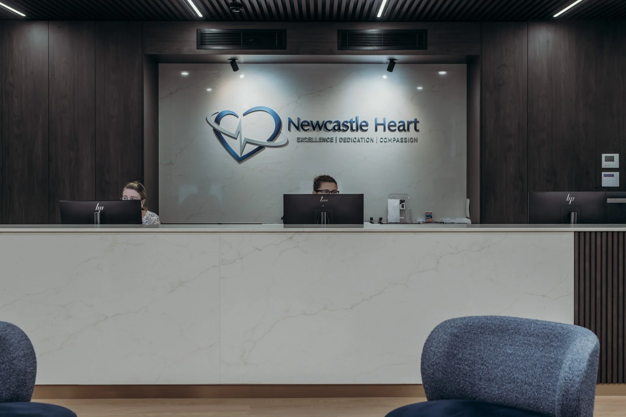 Reception area with a white marble counter, dark wood paneling, and a wall sign that says 'Newcastle Heart' with a heart logo and the words 'Excellence, Dedication, Compassion.' Two people are seated behind the counter using computers.