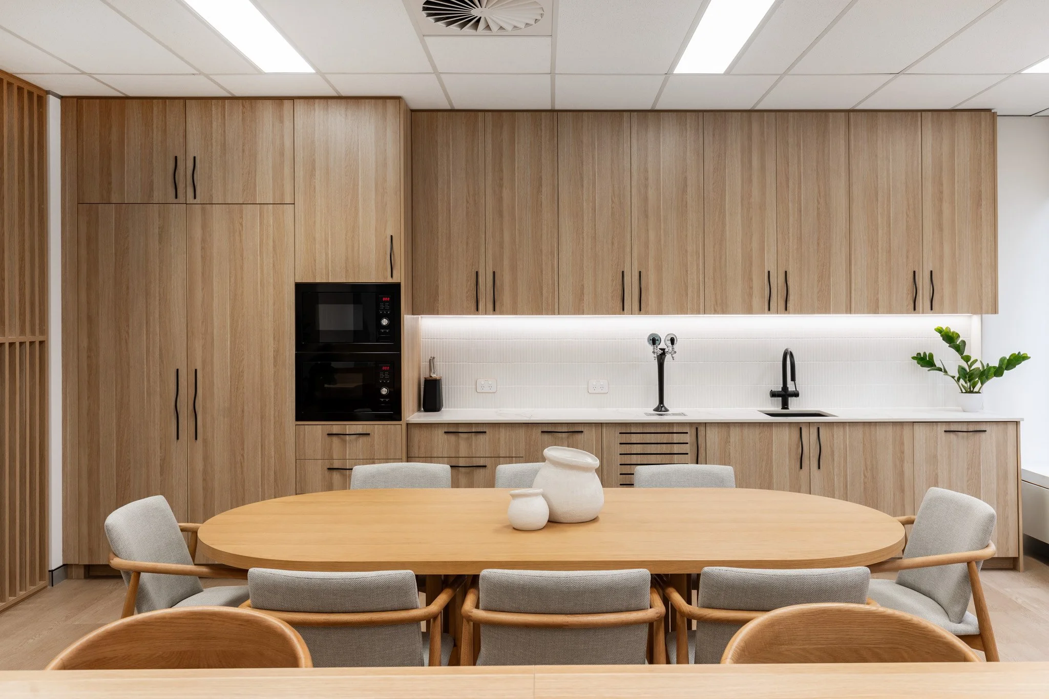 A modern kitchen and dining area with light wood cabinets, a round wooden table, and eight beige cushioned chairs. The kitchen has built-in black appliances, a white backsplash, a black sink, and a potted plant on the countertop. The ceiling has whit