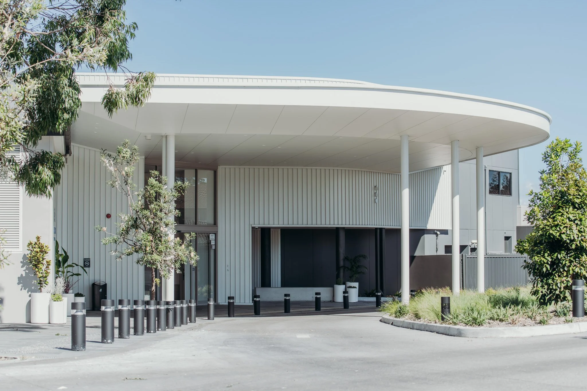 Modern white building with curved roof, surrounded by trees and small bushes, with black and gray bollards at the entrance.