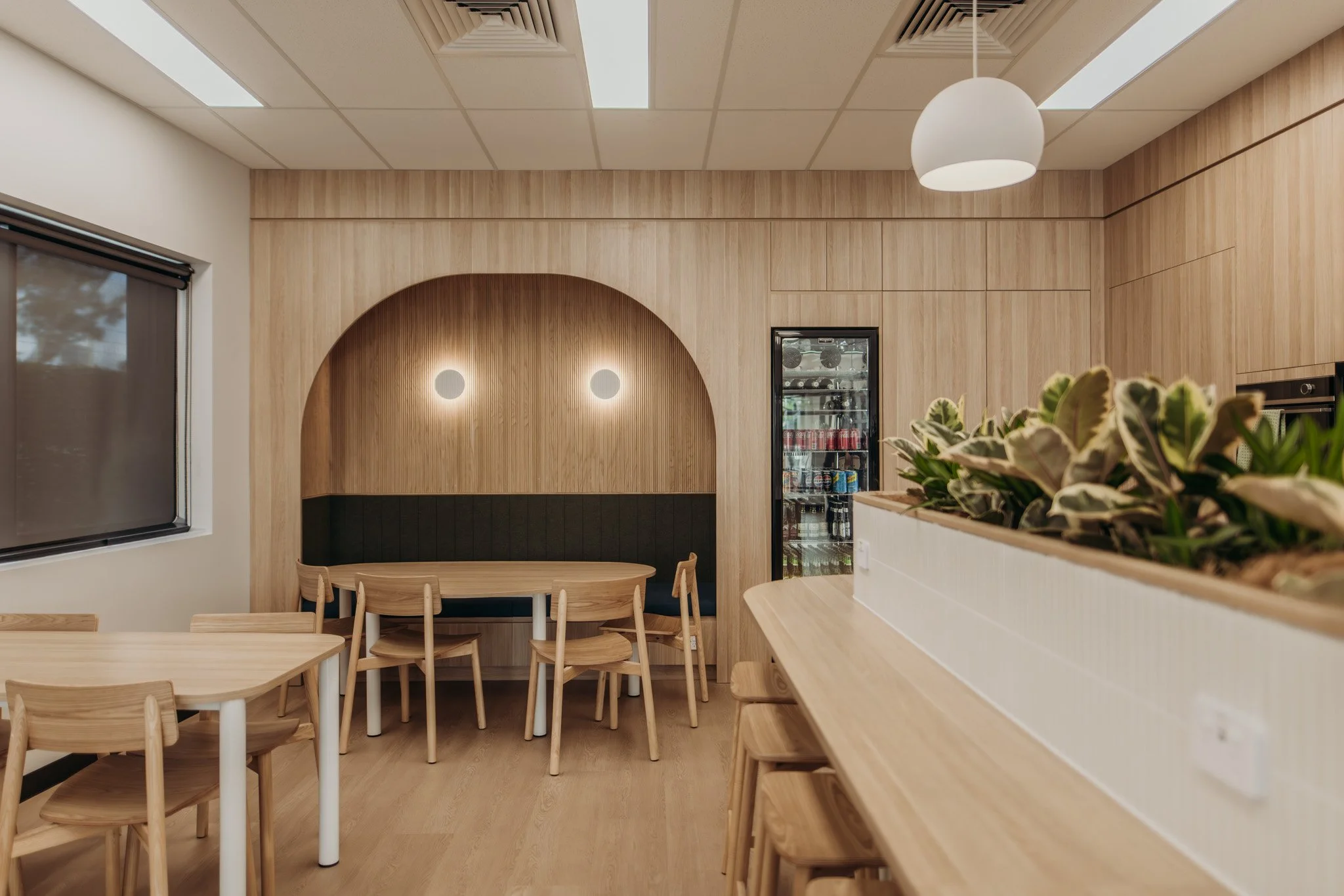 Modern cafe or restaurant interior with wooden tables and chairs, a wall with wooden paneling, a glass fridge with beverages, and a chandelier overhead.