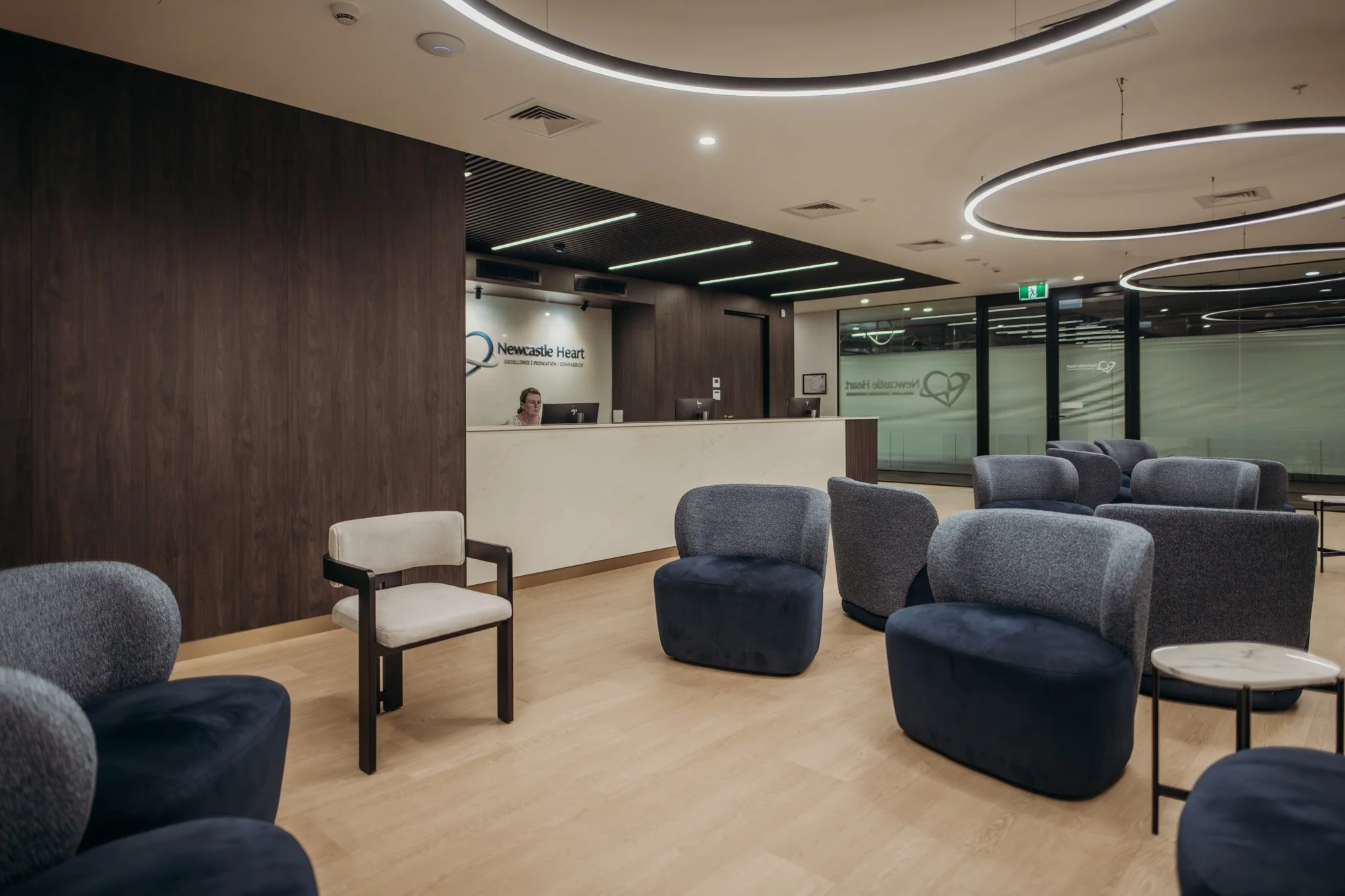 Interior of a modern medical reception area with a curved ceiling light, a reception desk with a person working, and seating with gray and navy upholstered chairs.