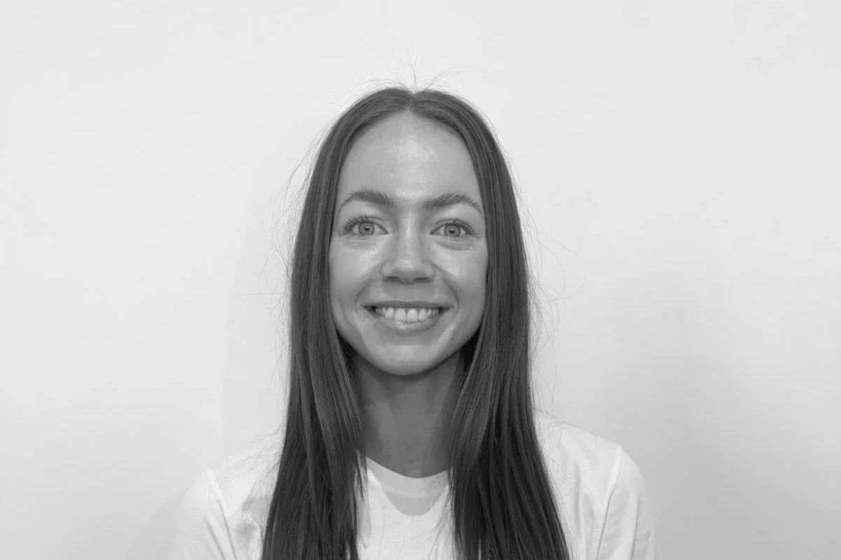 Black and white photo of a woman with long, straight hair smiling in front of a plain light-colored wall.