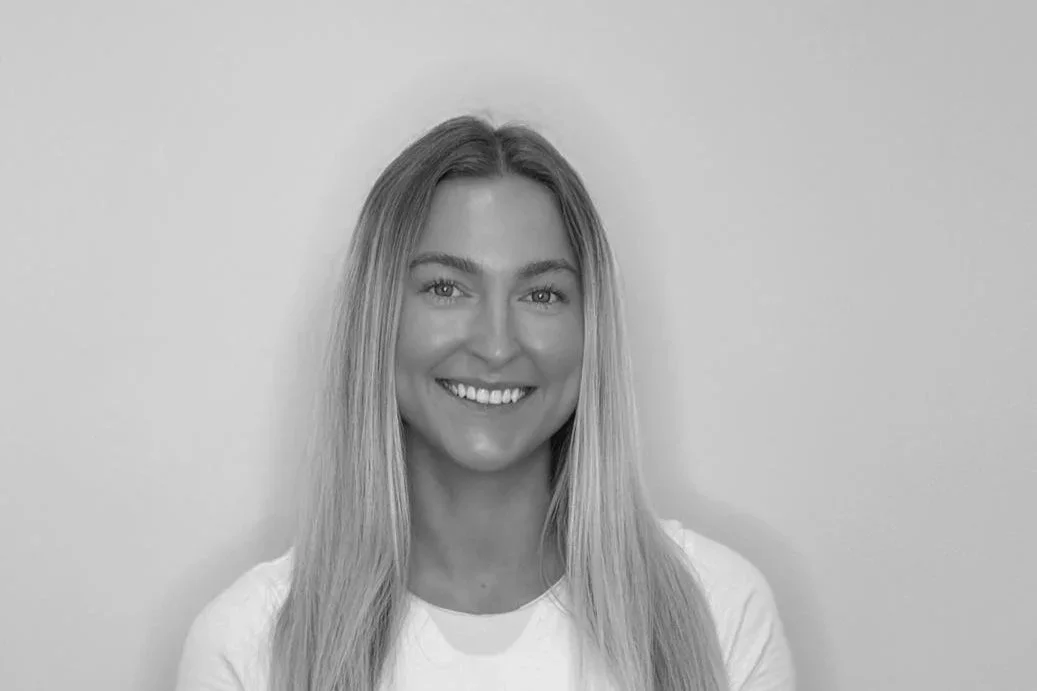 Black and white photo of a smiling woman with long straight hair, wearing a white shirt, standing against a plain wall.