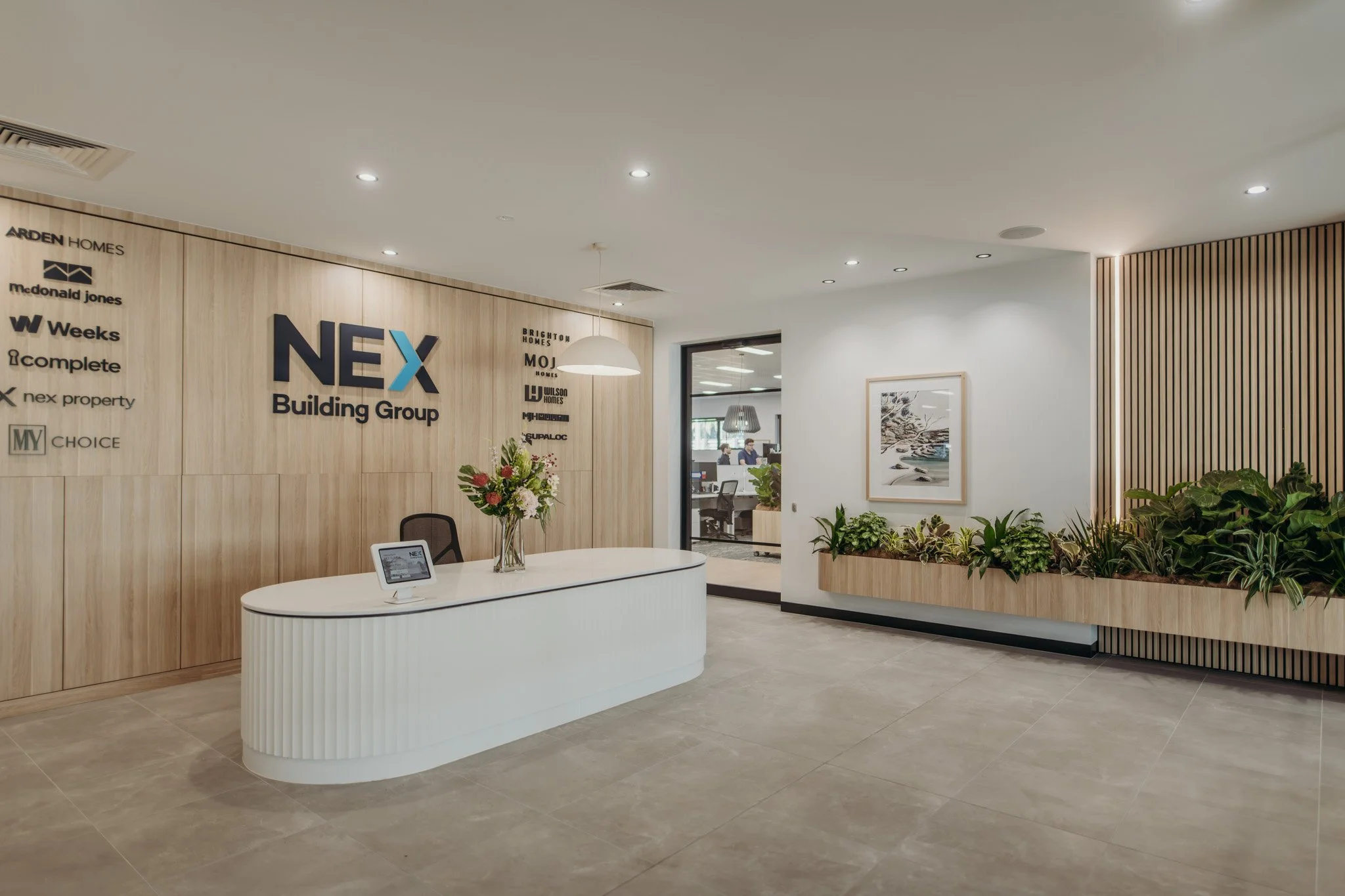 Modern office reception area with a curved white desk, wooden wall with company logos, a bouquet of flowers, and indoor plants, with a doorway leading to an office space in the background.