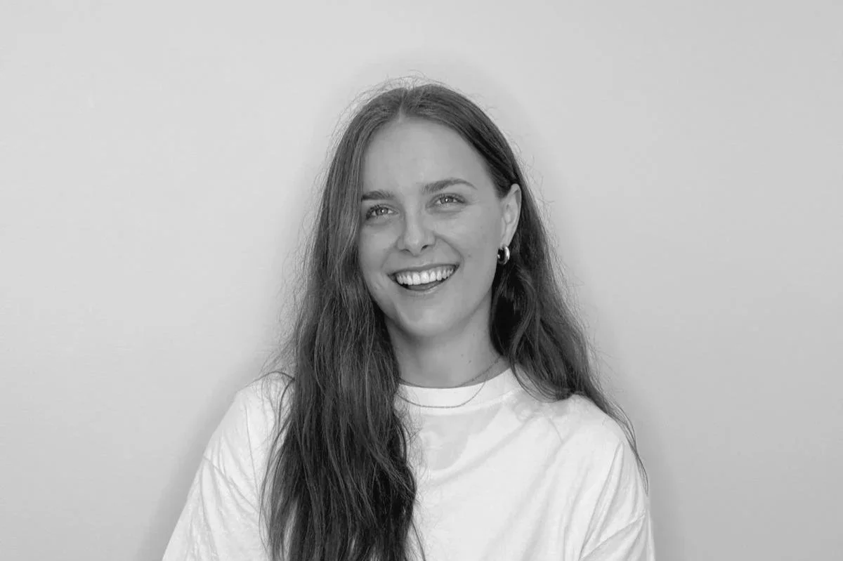 A young woman with long wavy hair, smiling and wearing a white t-shirt, standing against a plain wall.