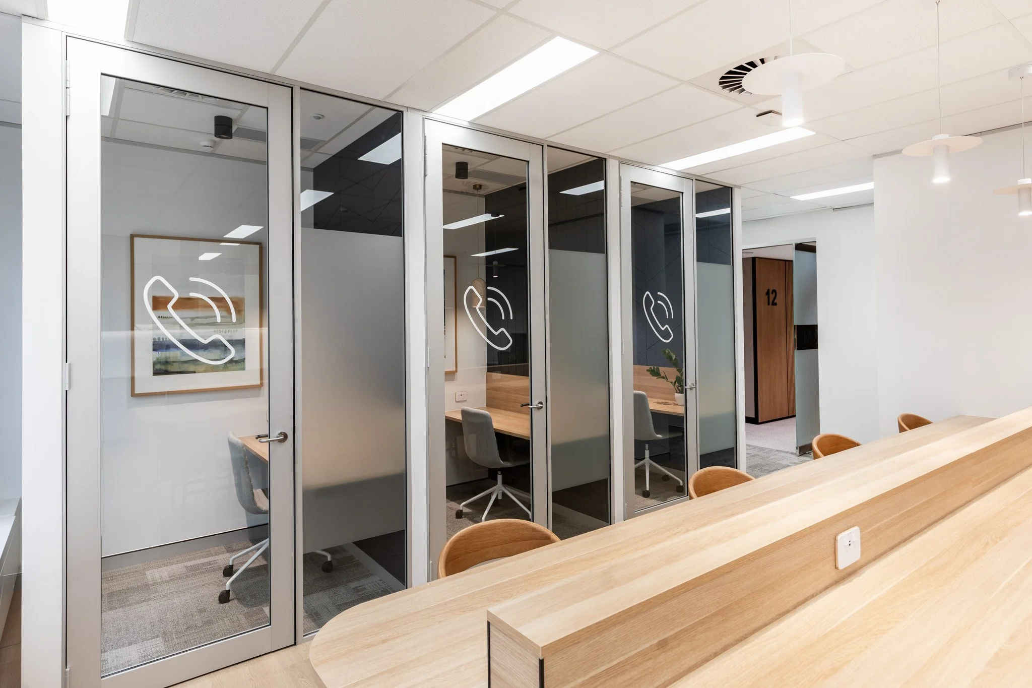 Modern office space with glass-walled meeting rooms, wooden tables, and chairs, illuminated by ceiling lights.