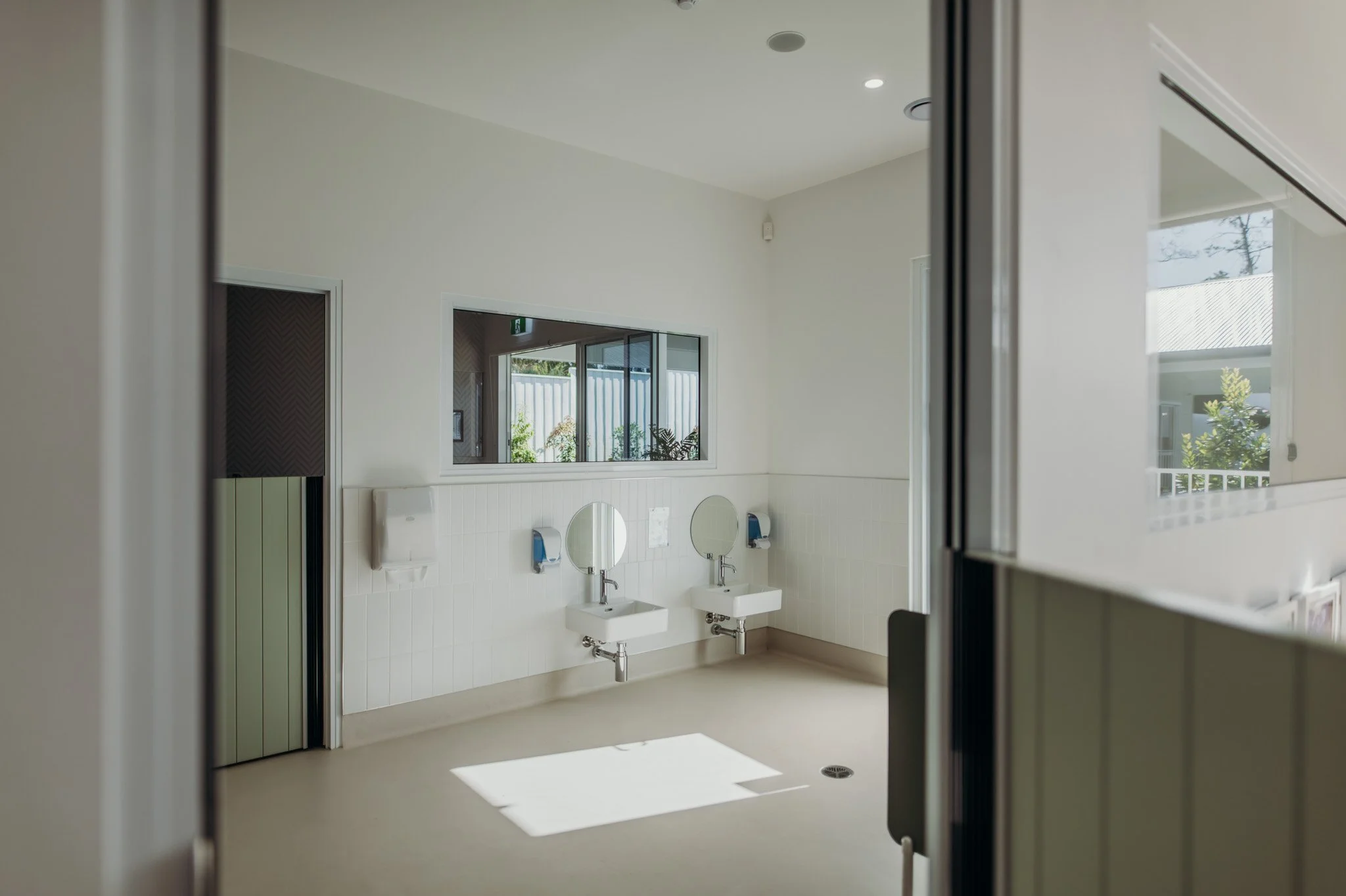 A public restroom with two sinks, mirrors, and soap dispensers, viewed through an open door with large windows showing outdoor greenery.