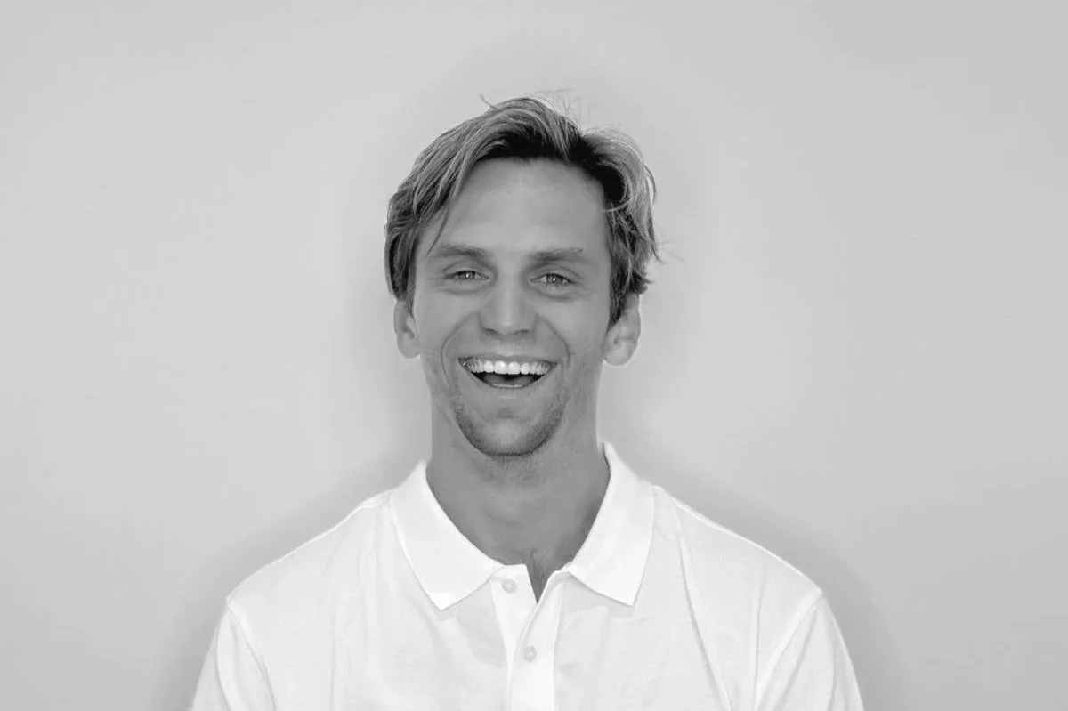 Black and white photo of a young man with light hair, smiling, wearing a white polo shirt, standing against a plain light background.