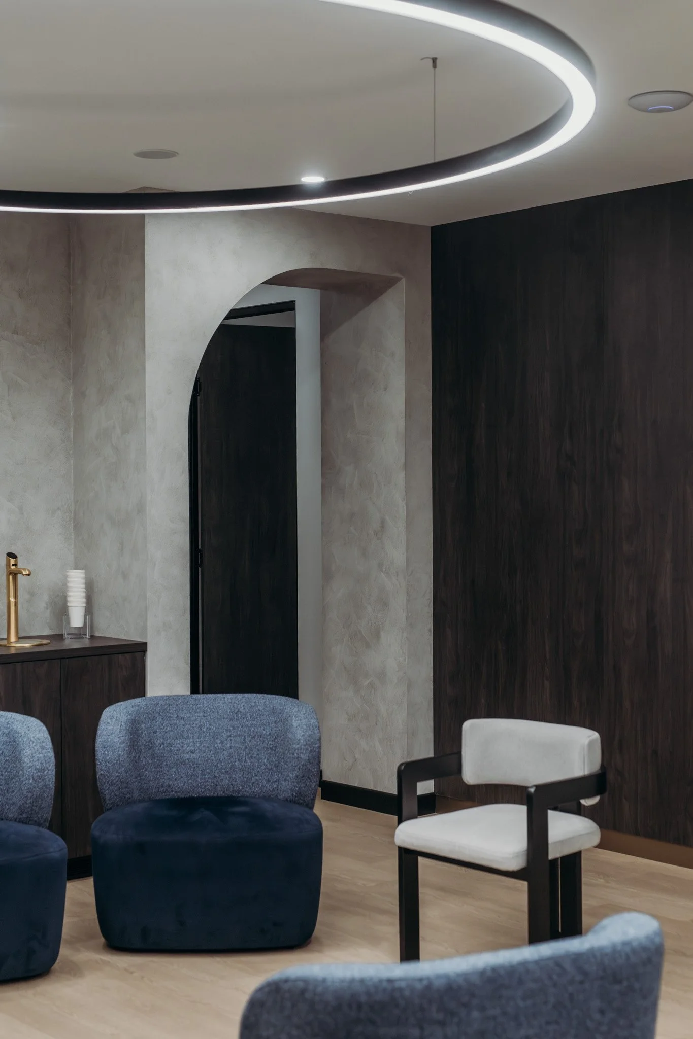 Modern interior room with dark wood and textured gray walls, round ceiling light fixture, and Various chairs including blue velvet ottomans and a white armchair.