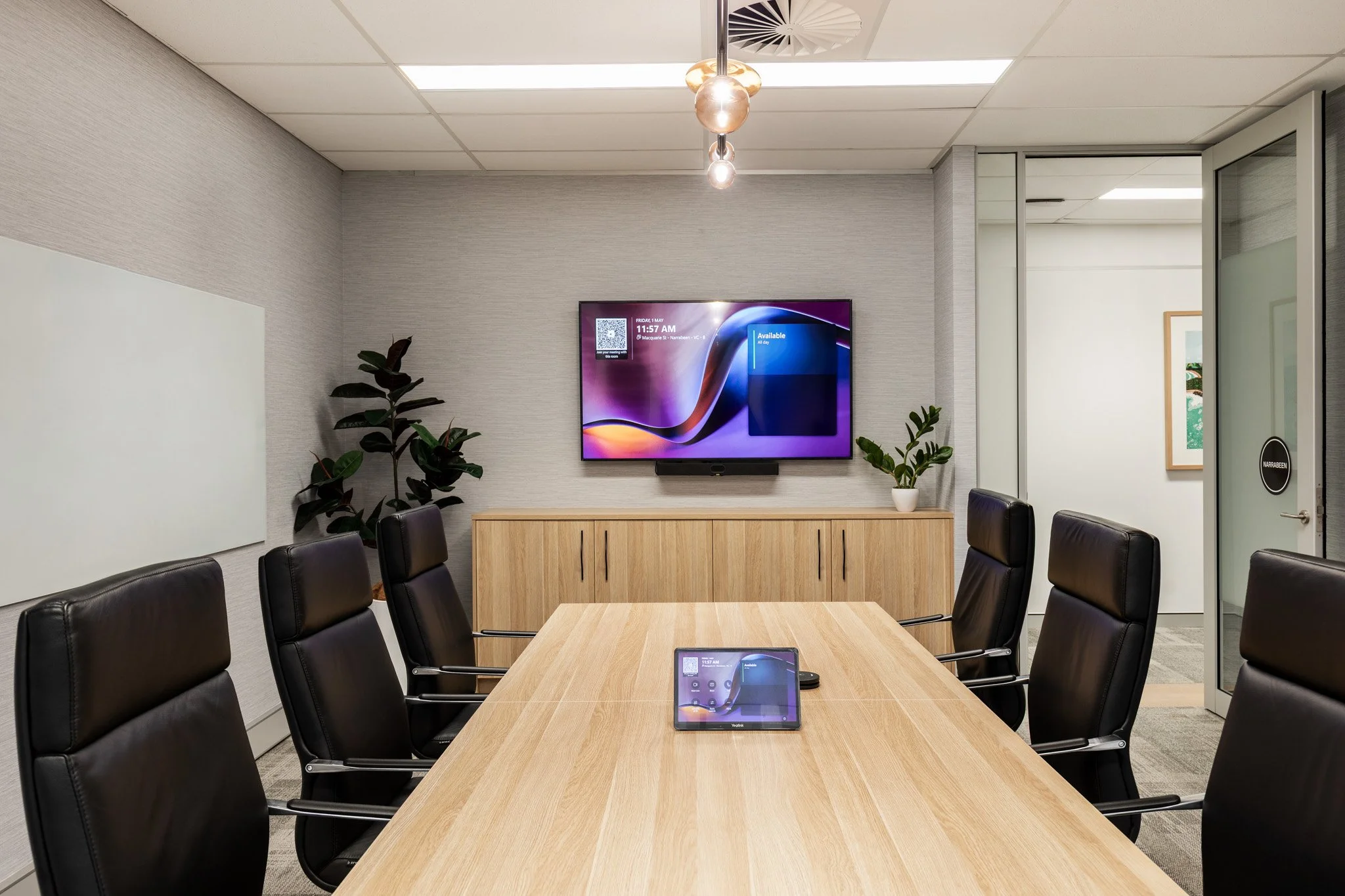 Conference room with a wooden table, black chairs, a wall-mounted TV displaying a screen, and some plants.