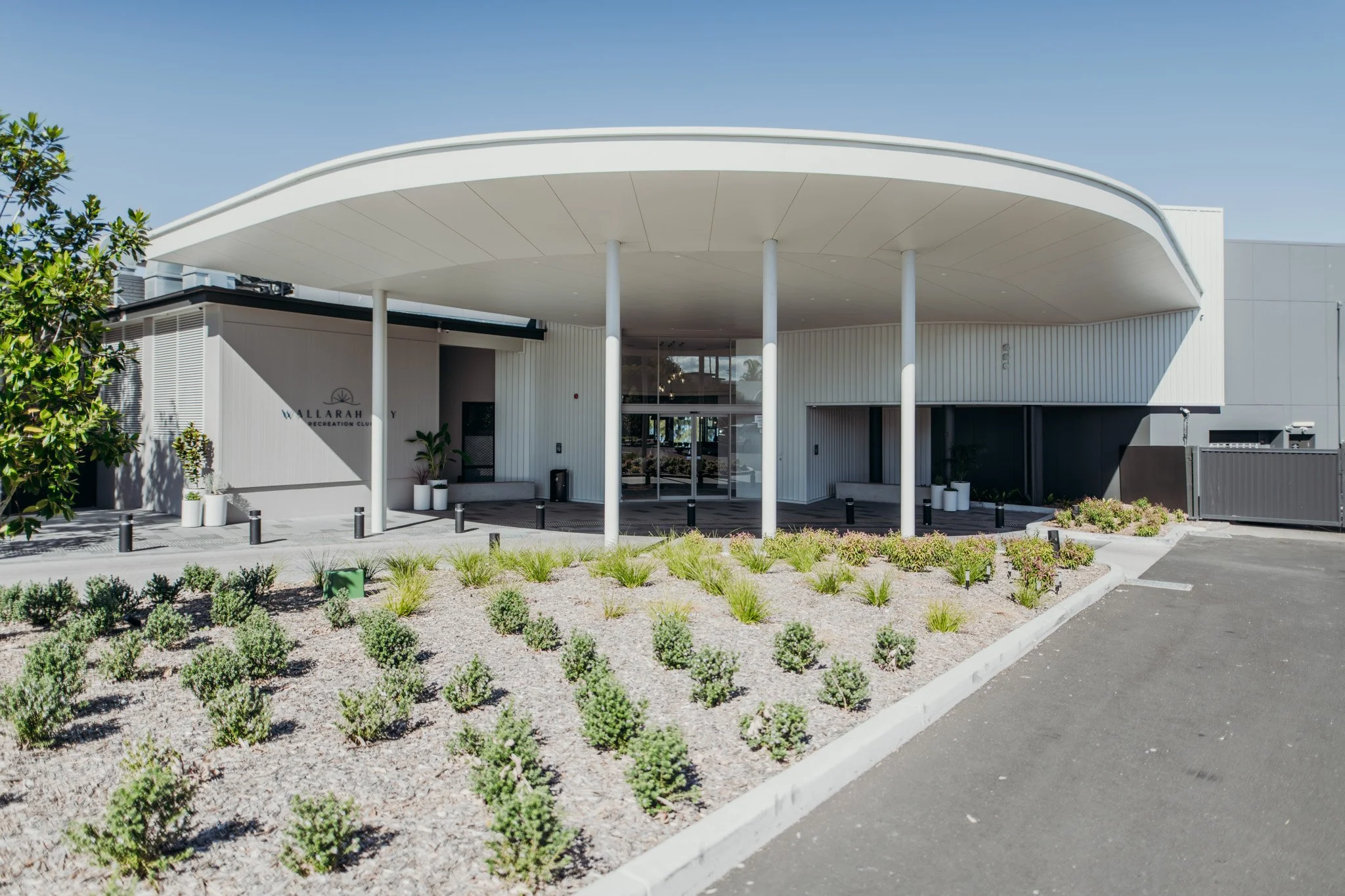 Exterior view of the Wallarah Youth Recreation Club building with a modern design featuring a curved white roof supported by slender white columns, surrounded by landscaped greenery and a parking area.