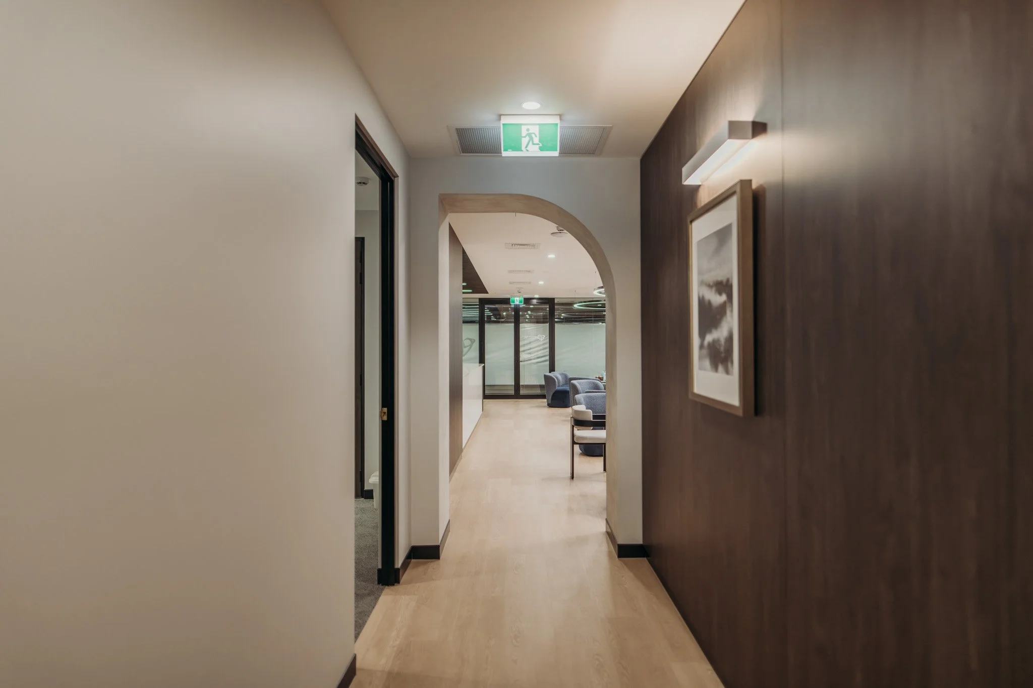 Hallway with beige walls, wood-paneled accent wall on the right, modern framed art, and seating area at the end with chairs and a glass door.
