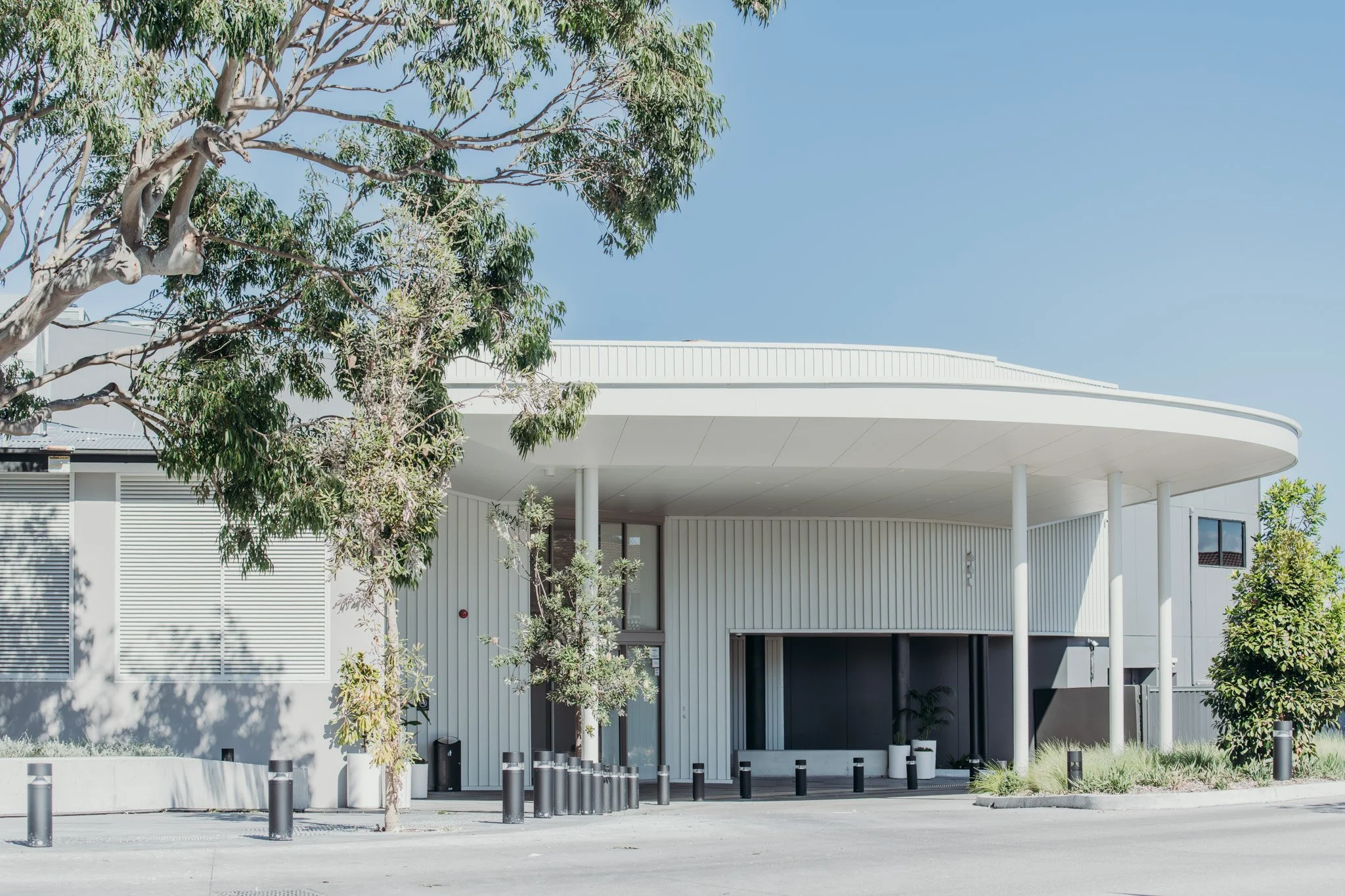 Modern white building with curved roof, surrounded by trees and small plants, situated in a paved area with bollards.