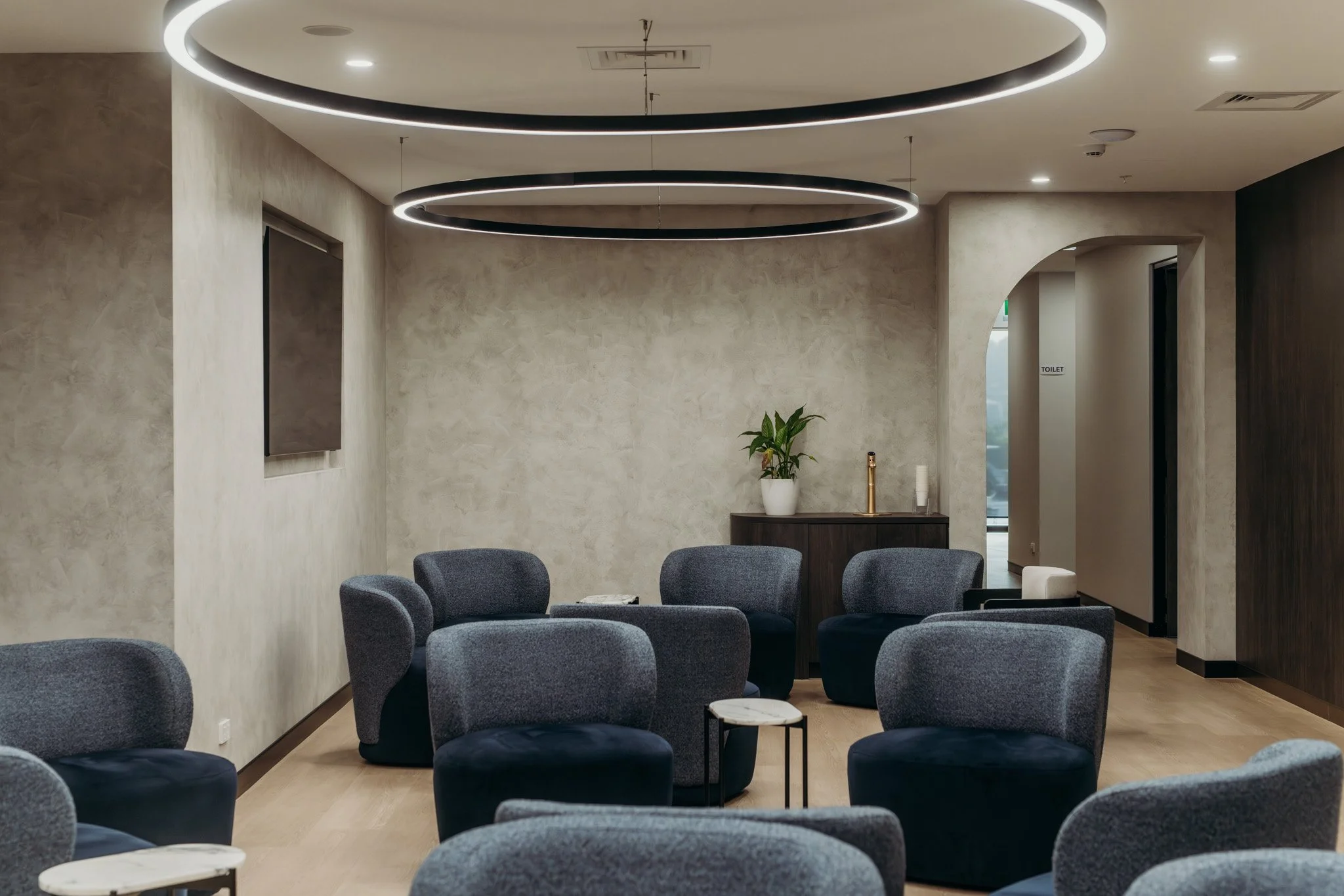 A modern waiting room with dark gray chairs arranged around small side tables, beige textured walls, a plant on a dark wooden table, and circular ceiling lights.