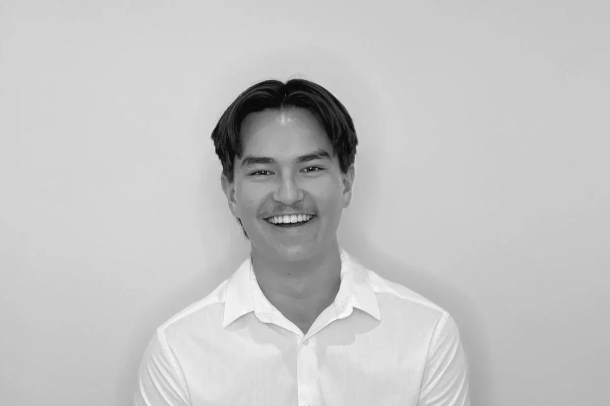 A black-and-white portrait of a young man with dark hair, smiling, wearing a white collared shirt, standing against a plain light-colored background.